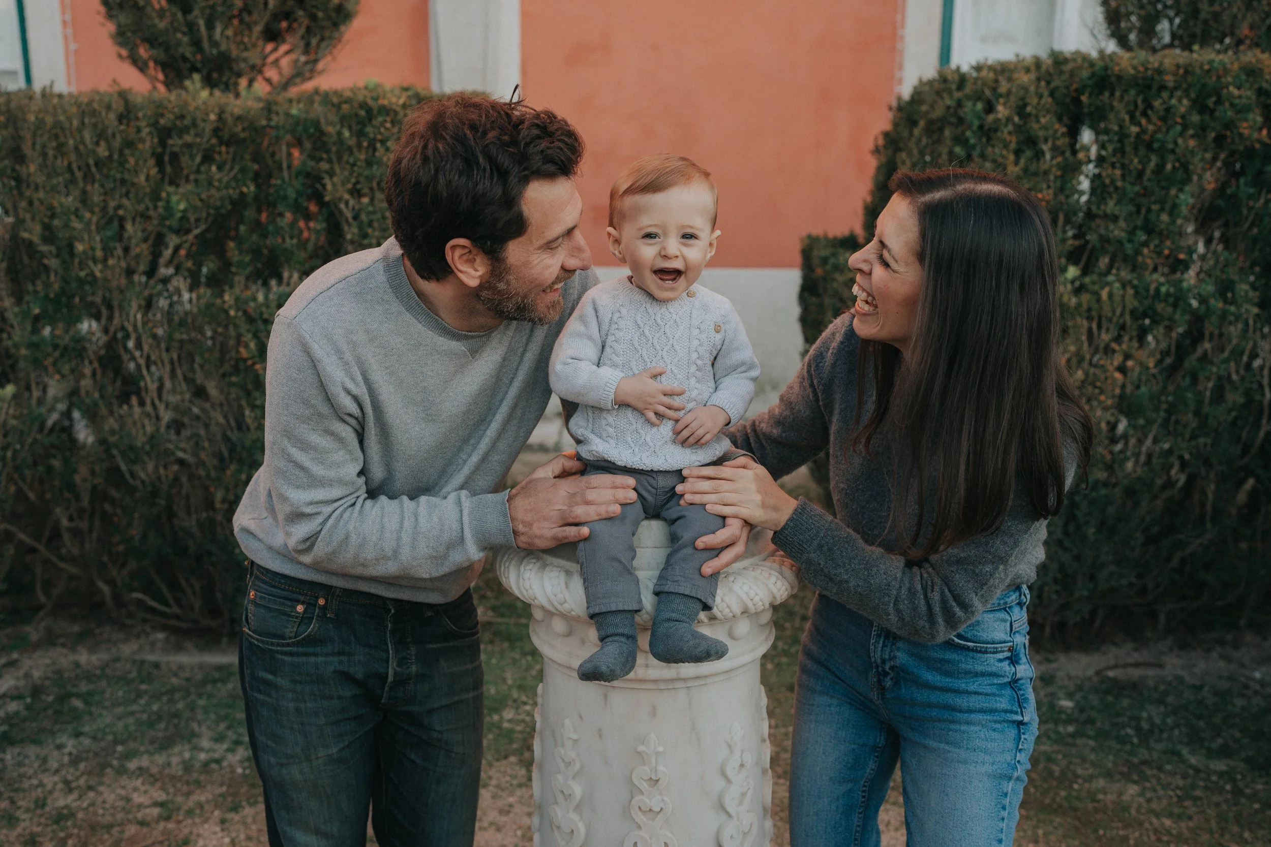 A young child sitting on a decorated white pedestal outdoors, smiling and crying while being held by a man and woman. The man has dark hair and a beard, wearing a gray sweater, and the woman has long dark hair, wearing a gray sweater. They are sharin