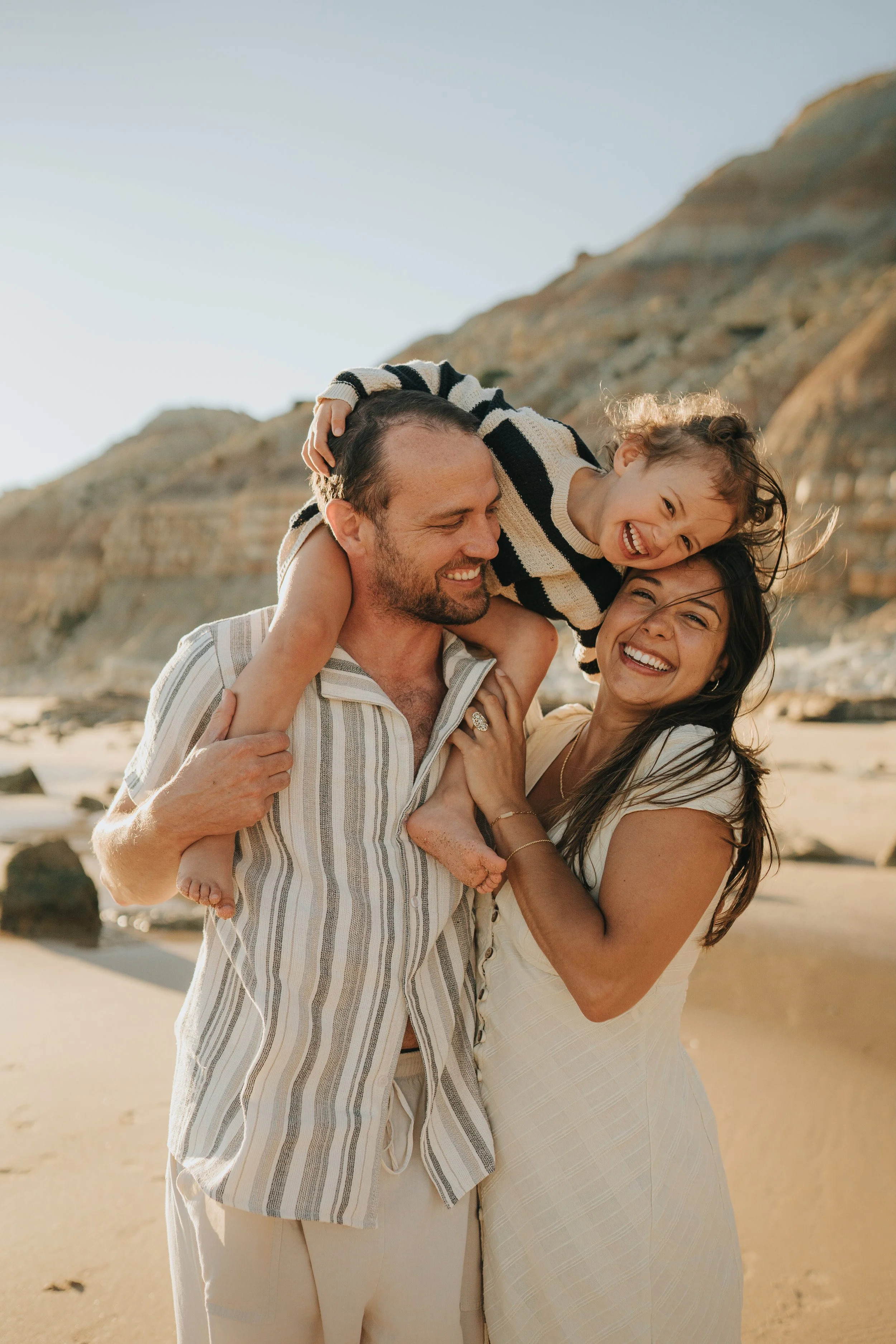 Family of three enjoying a moment on the beach with a rocky hillside in the background, smiling and having fun.