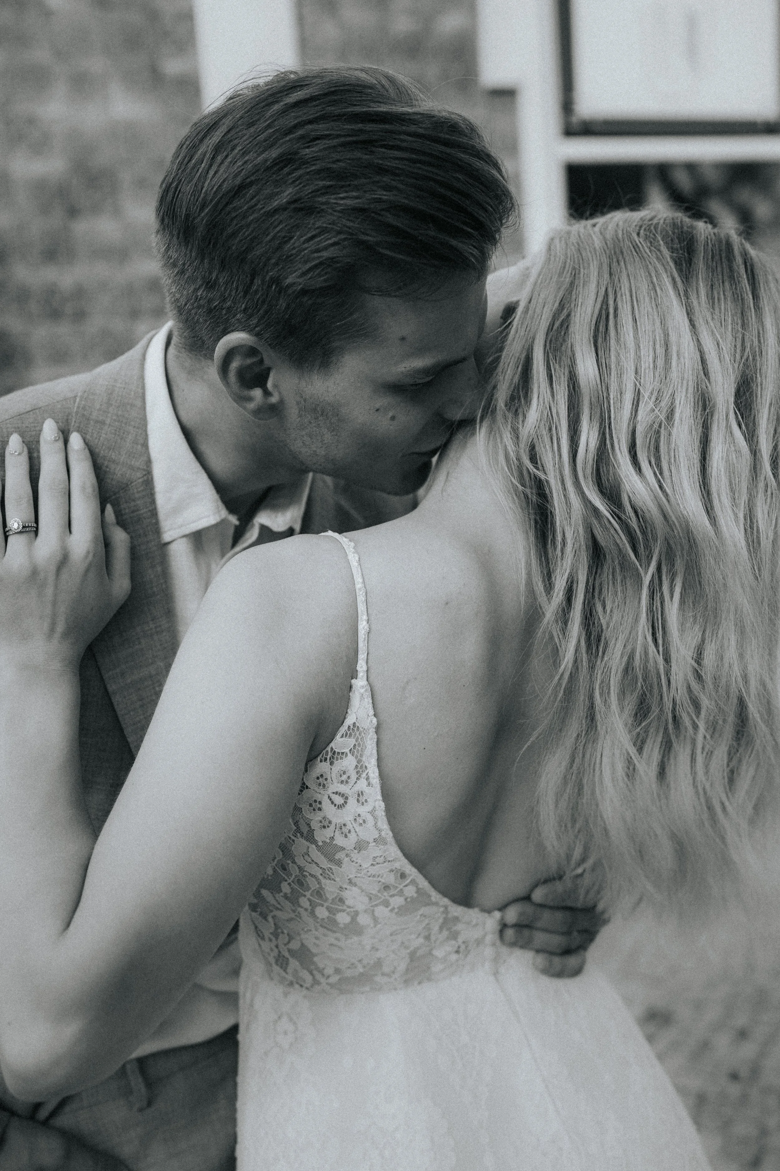 A black and white photo of a man and a woman kissing. The woman's hand with a ring rests on the man's shoulder. The woman has long, wavy hair and is wearing a lace dress. The man has short, neatly styled hair and is dressed in a suit.