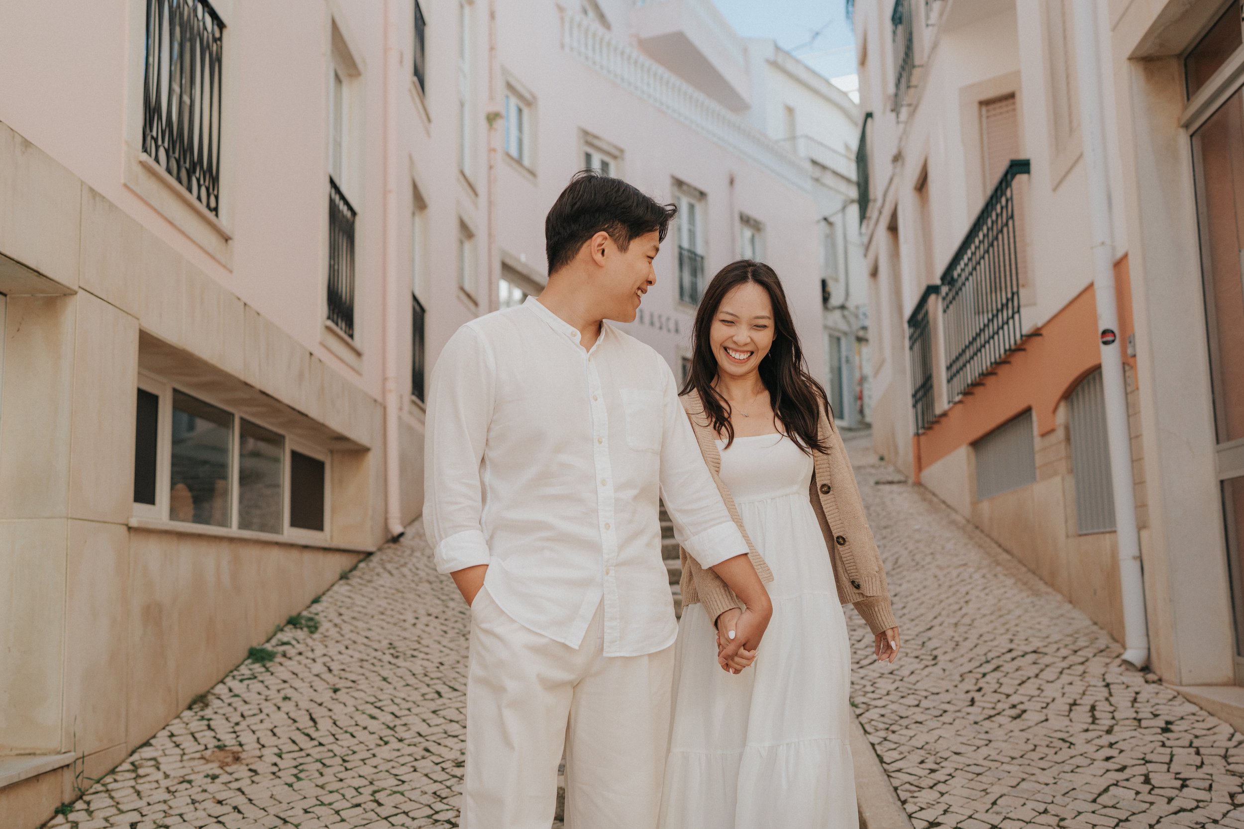 A young couple walking hand in hand down a cobblestone street, smiling and looking at each other, in a European-style neighborhood with pastel-colored buildings and balconies.