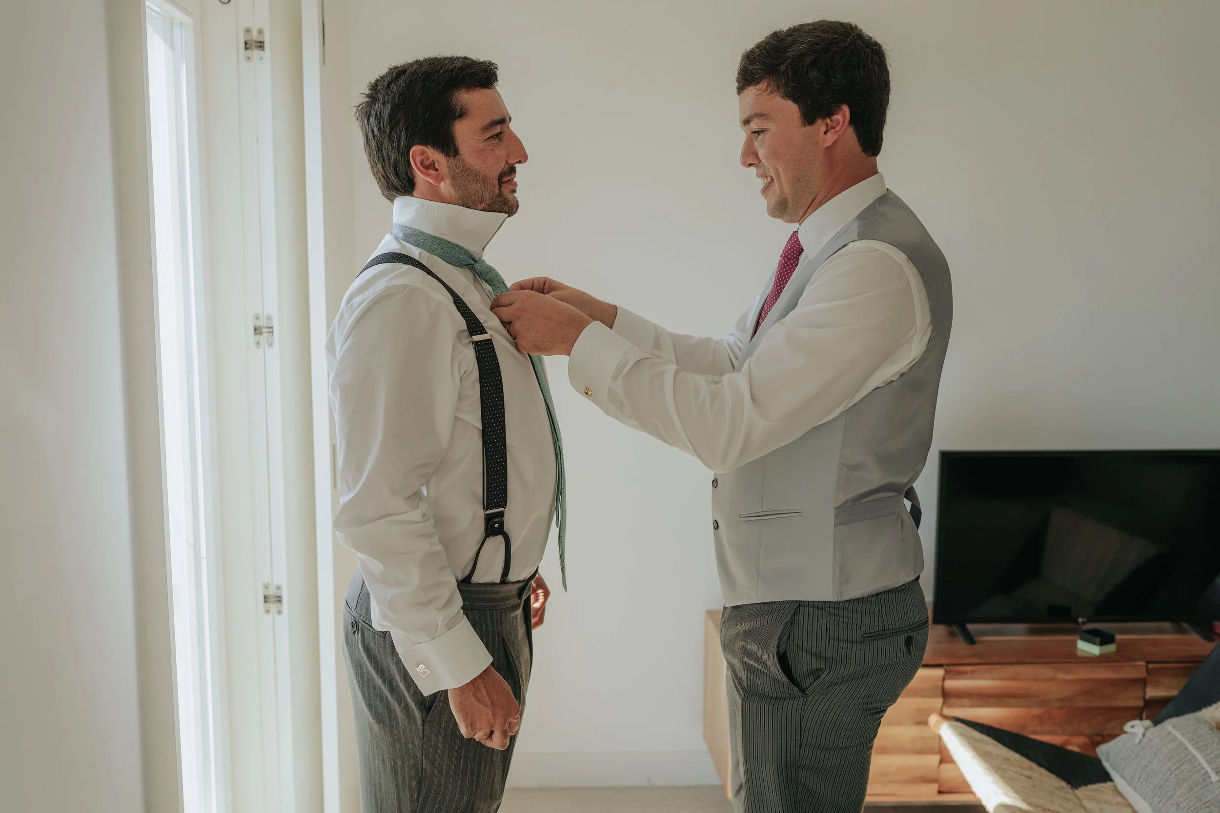 Two men in suits are in a living room, with one adjusting the other's tie, indicating a moment of preparation or celebration.