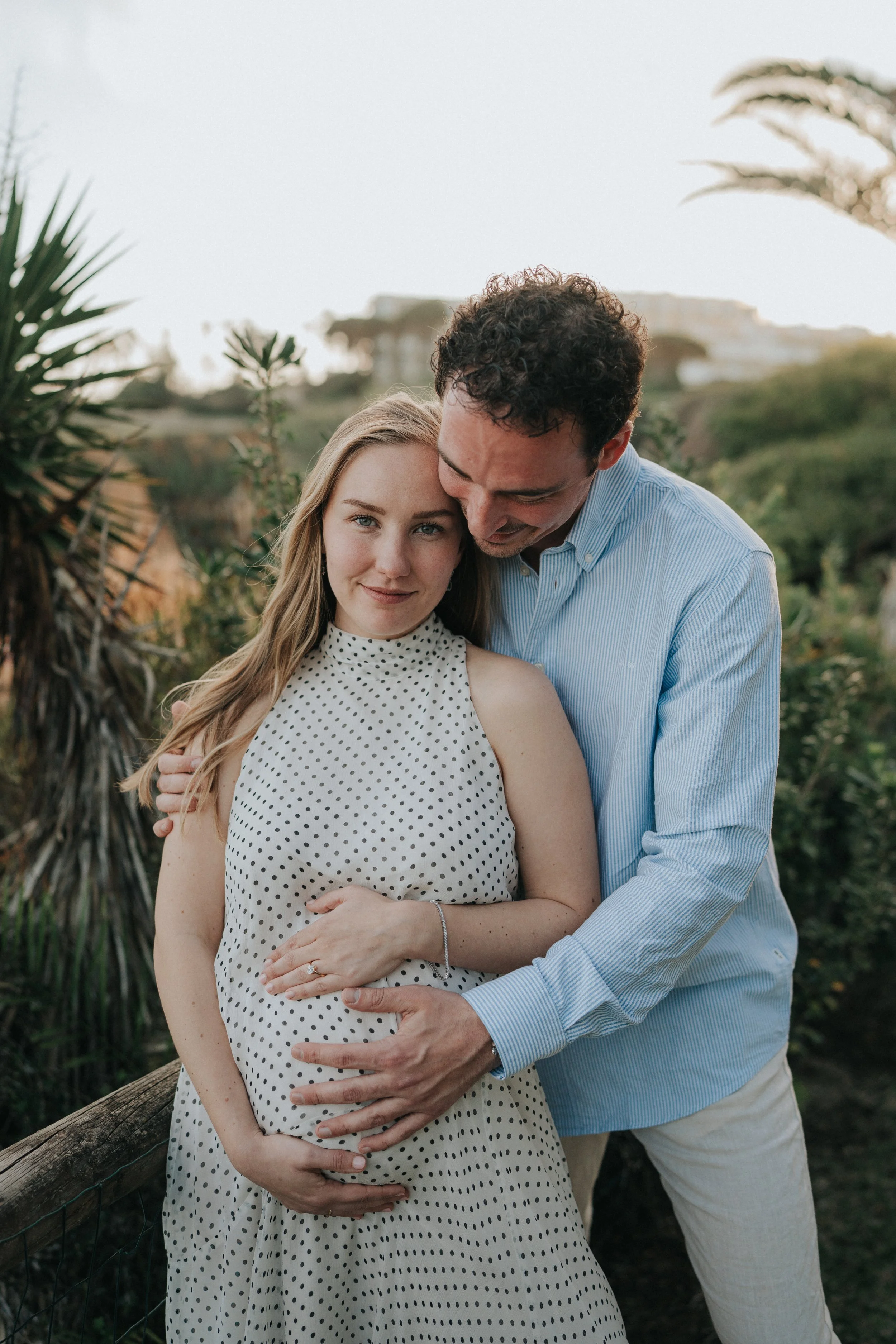 A pregnant woman wearing a white dress with black polka dots and a man in a light blue shirt with white pants, standing outdoors with greenery and trees in the background. The man is embracing the woman and both are smiling gently, with the woman loo