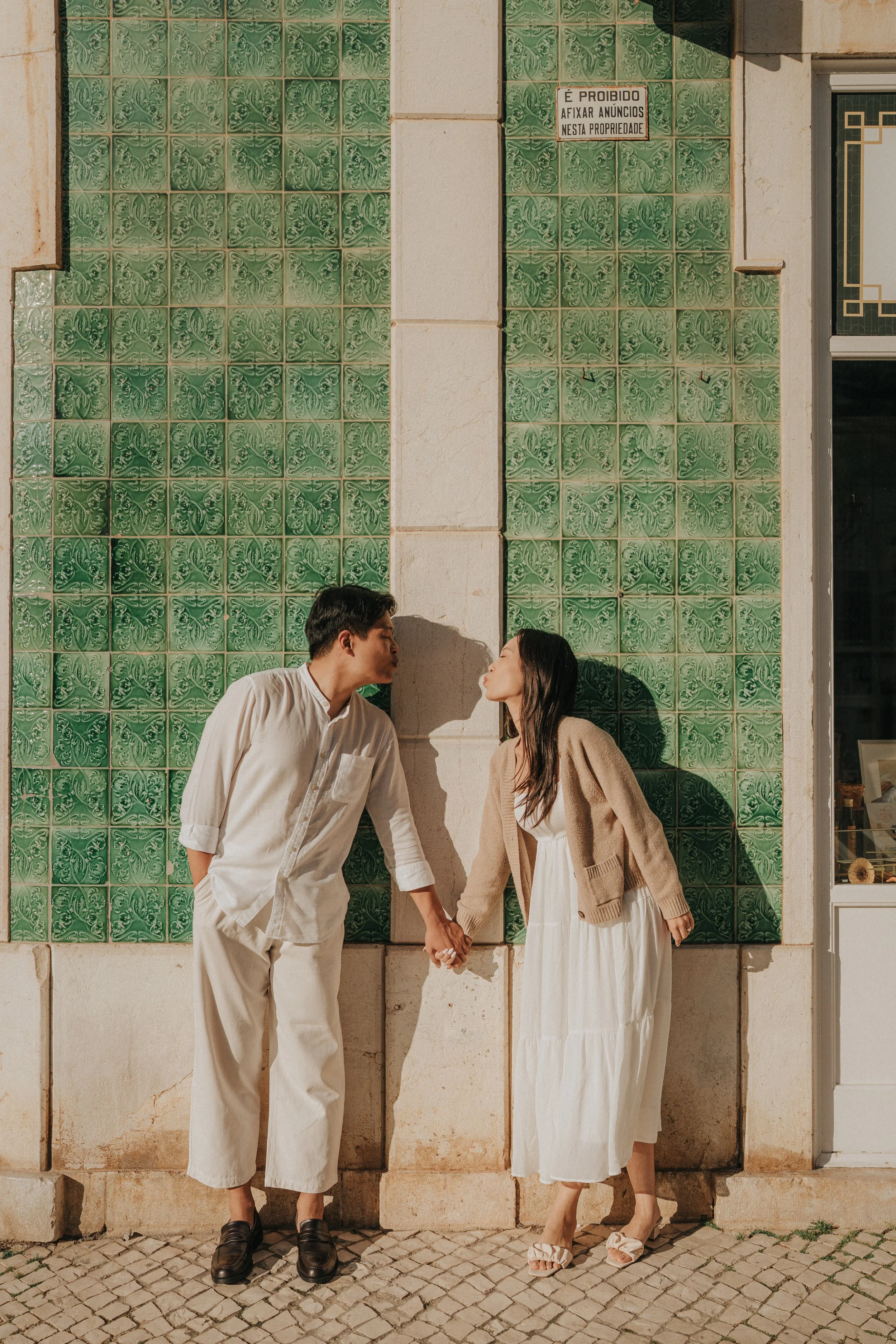 A couple is standing outdoors, holding hands, facing each other and about to kiss. They are standing in front of a wall with green decorative tiles, and the man is wearing a white shirt and pants while the woman is in a beige cardigan and a white dre