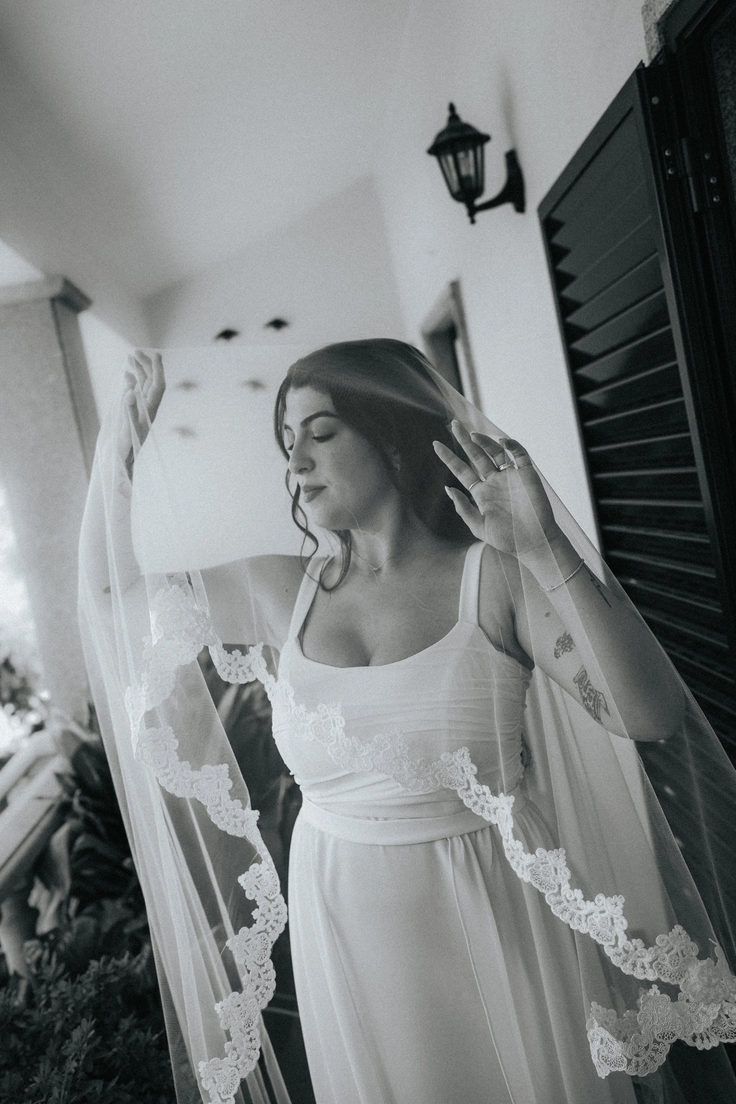 A woman wearing a wedding dress and veil, holding up the veil with one hand, standing indoors near a wall with lantern-style light fixtures and a window.