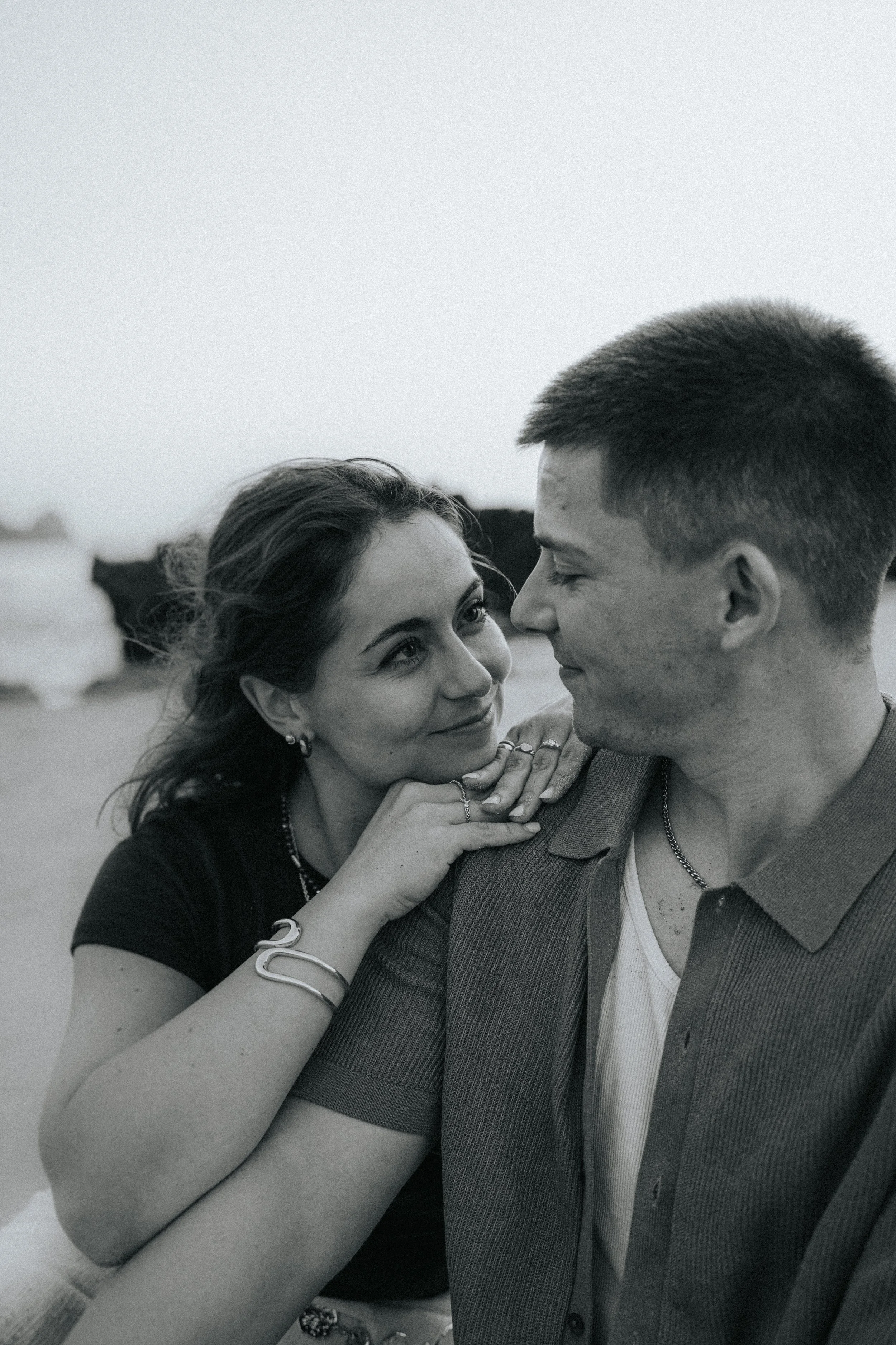 A black and white photo of a couple standing close together, with the woman looking up at the man and the man looking down at her. The woman has dark, wavy hair and is wearing earrings, rings, and a bracelet. The man has short hair and is wearing a c