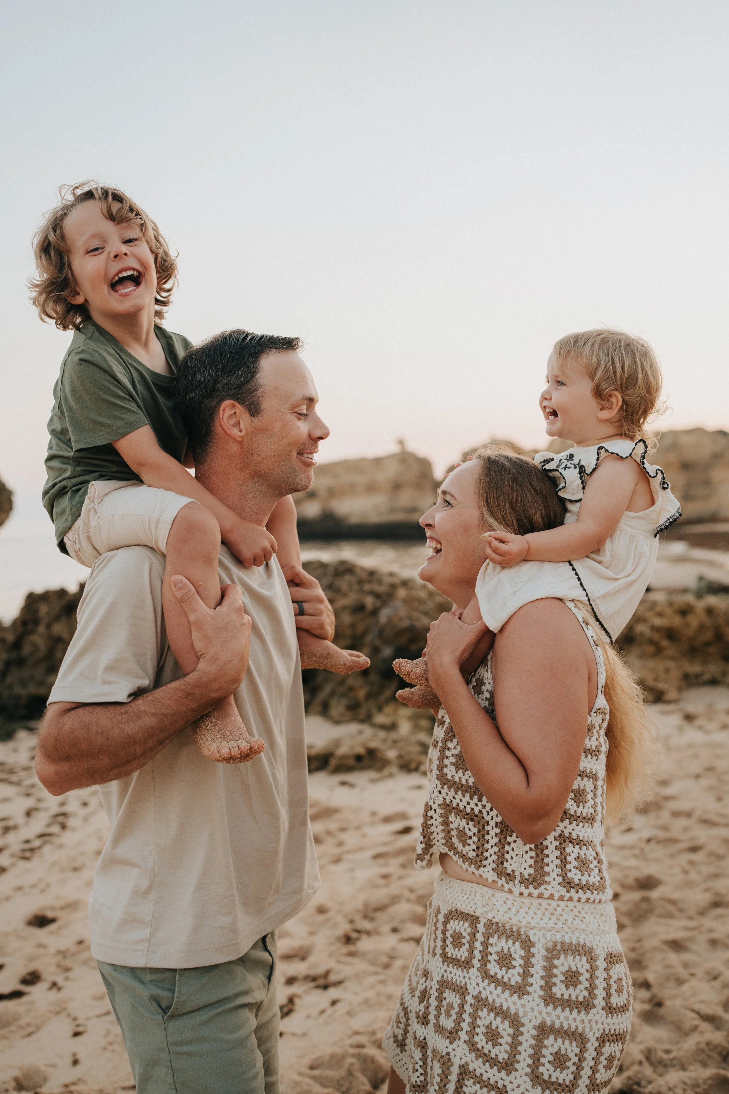 A family of four, consisting of a man, woman, and two children, enjoying a day at the beach during sunset, with rocky formations in the background.