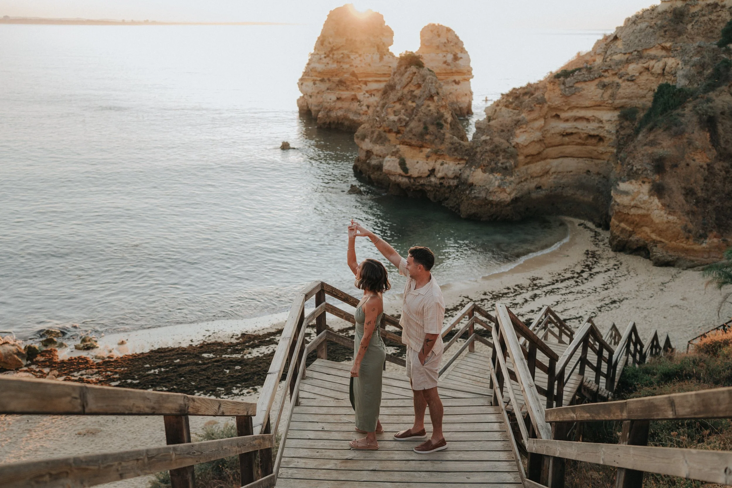 Three people standing on a wooden staircase at the beach during sunset. One woman and one man are looking at each other, and the woman is taking a photo with her phone. The background features large rock formations and calm ocean water.