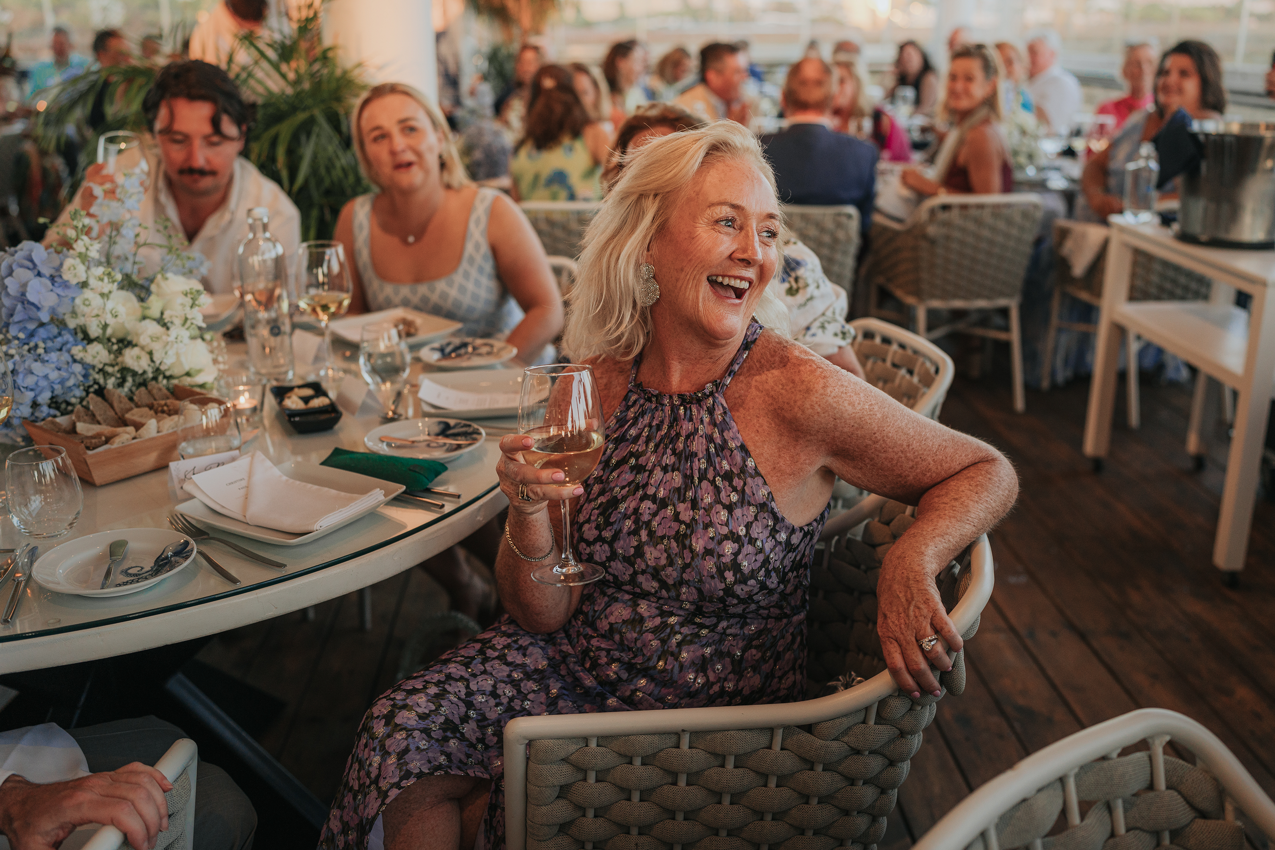 Group of people at a social gathering or celebration, sitting at a decorated table with food and drinks, including a woman in a patterned dress holding a glass of wine, smiling and enjoying the event.