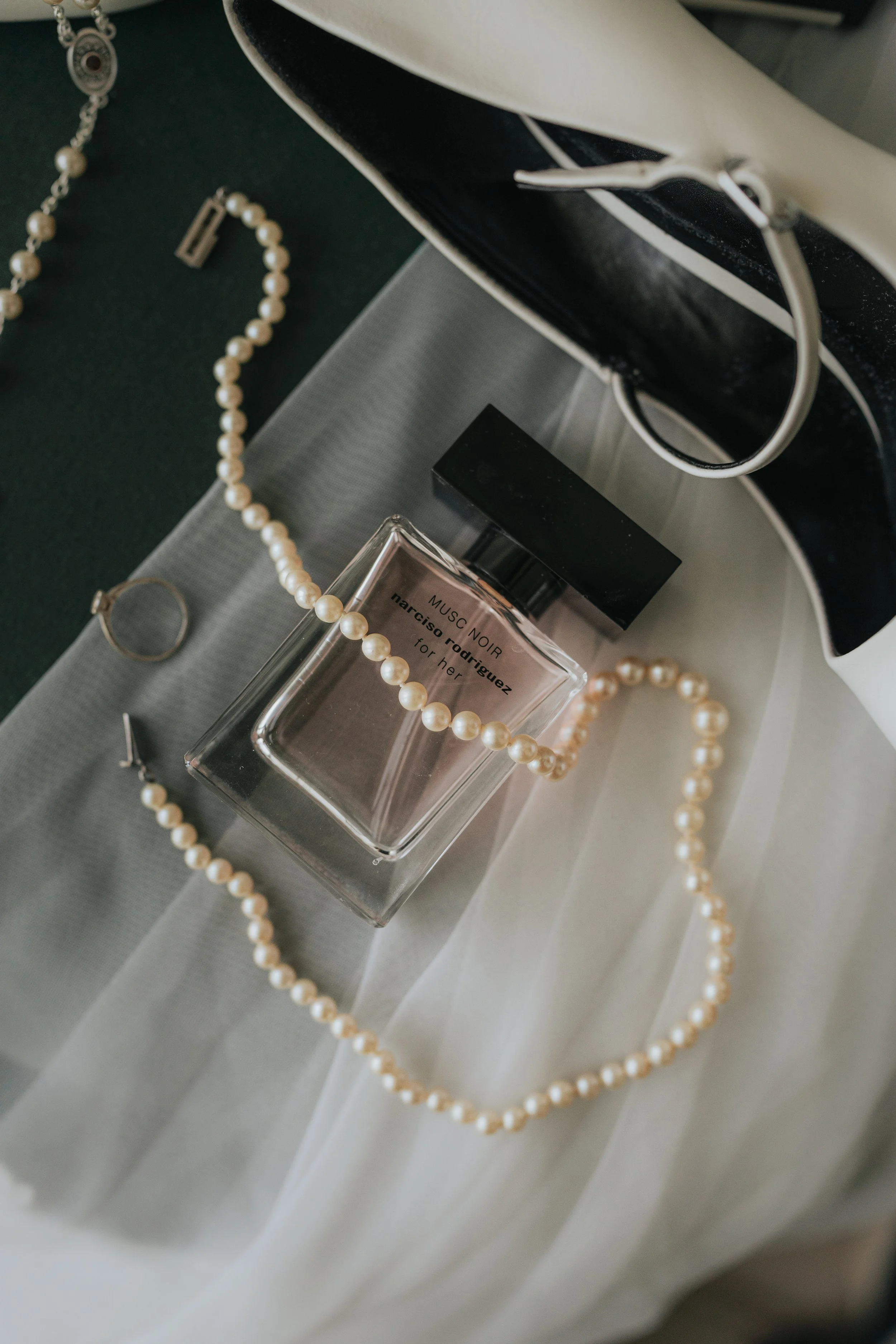 A perfume bottle labeled "MUSC NOIR" by Narciso Rodriguez, surrounded by a pearl necklace, a ring, and a bracelet, on a fabric surface.