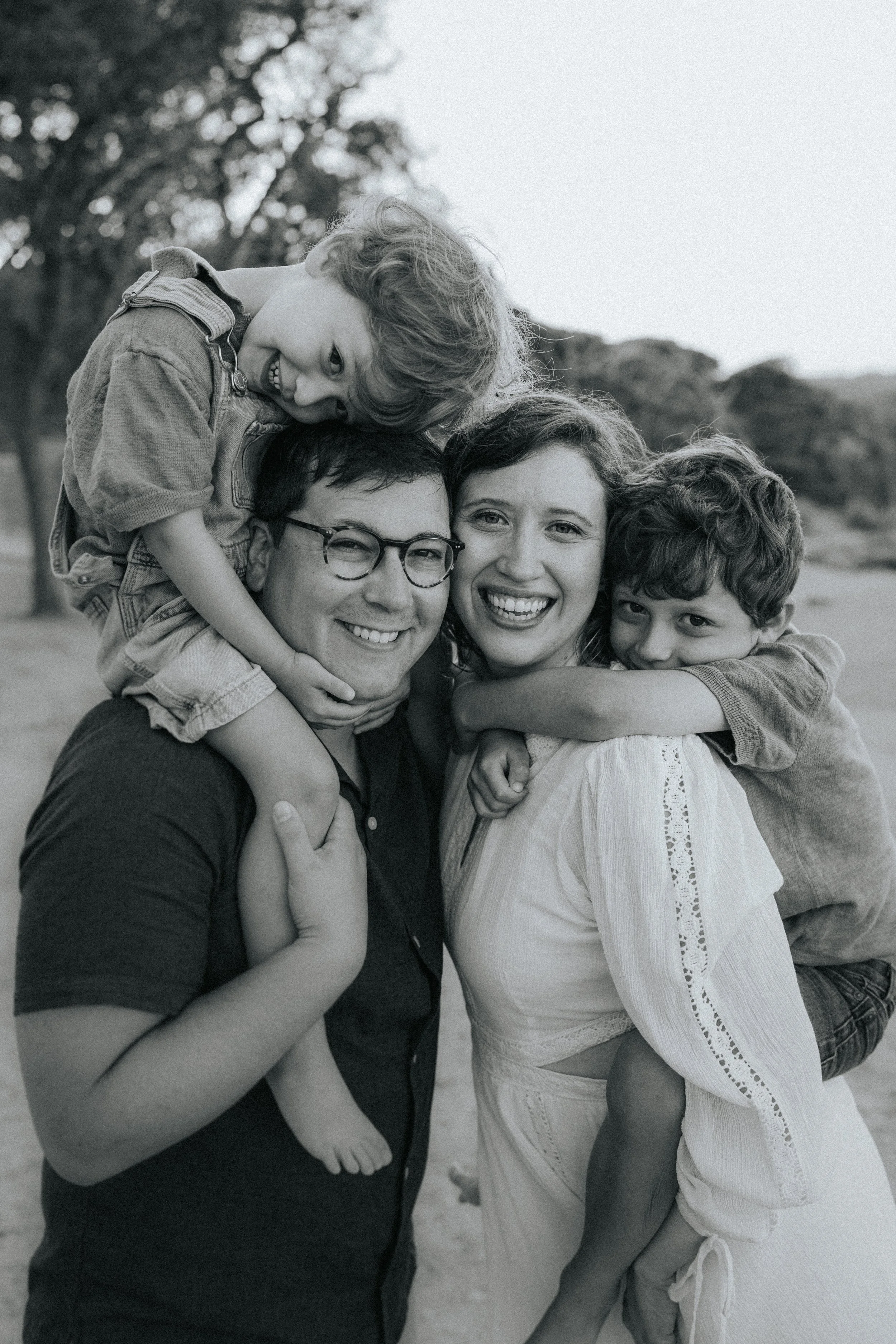 A happy family group consisting of two adults and two children hugging and smiling outdoors. The children are on the adults' shoulders and hugging them, with trees visible in the background.