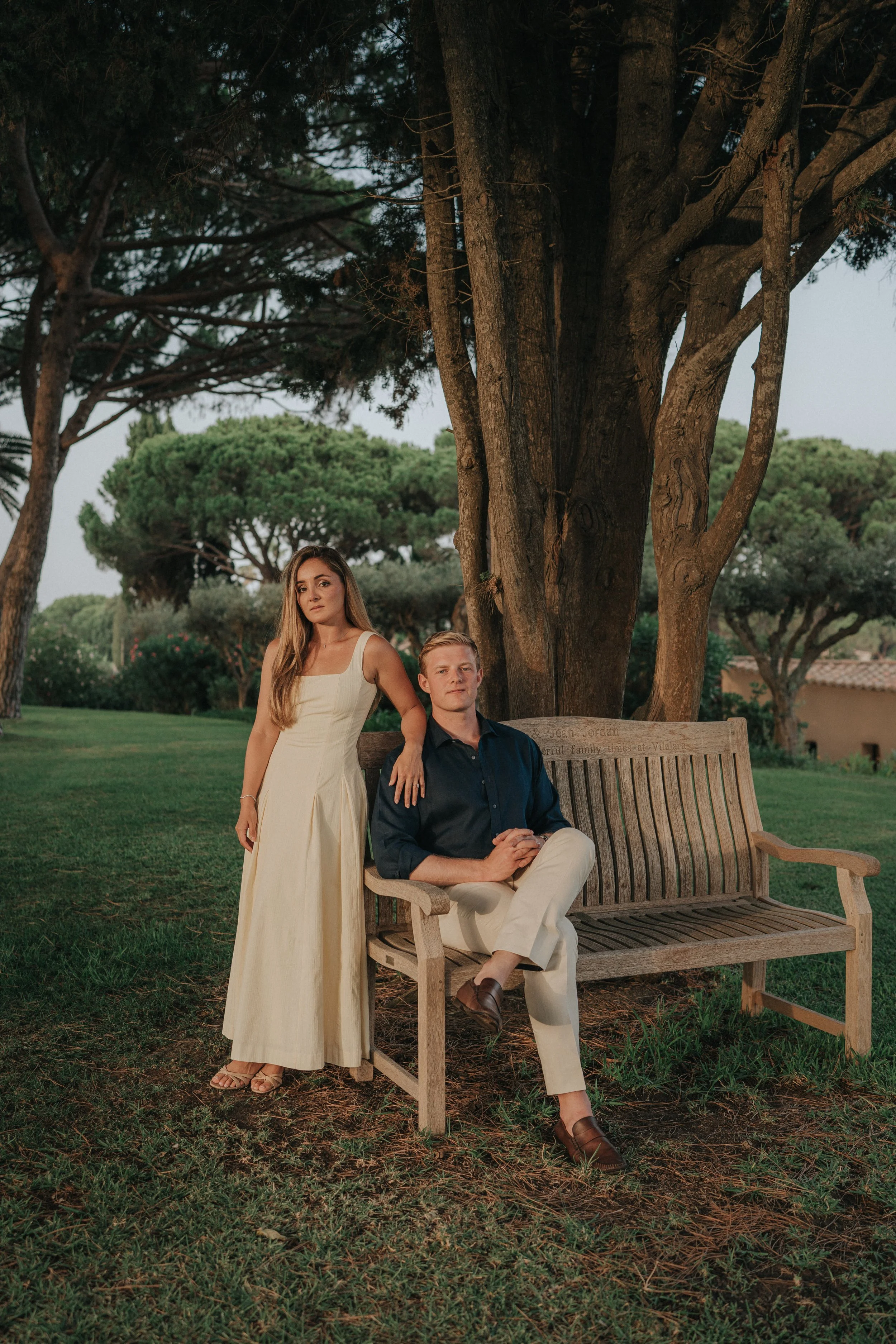 A woman in a long cream-colored dress standing next to a seated man in a dark shirt and light beige pants on a wooden bench outdoors under large trees during dusk.