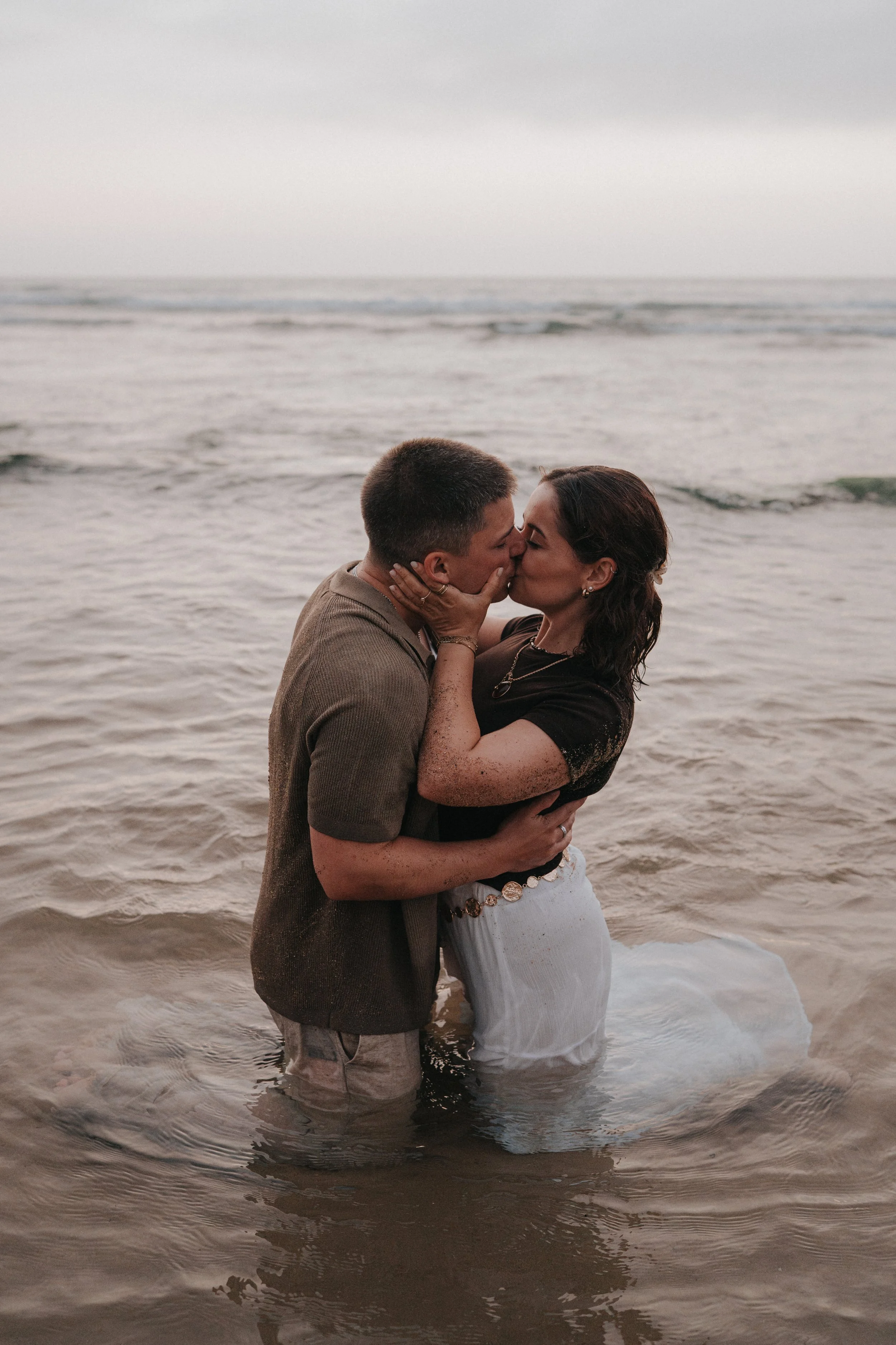 A couple shares a kiss while standing in the ocean at the beach during sunset, with their feet in the water and waves gently lapping around them.