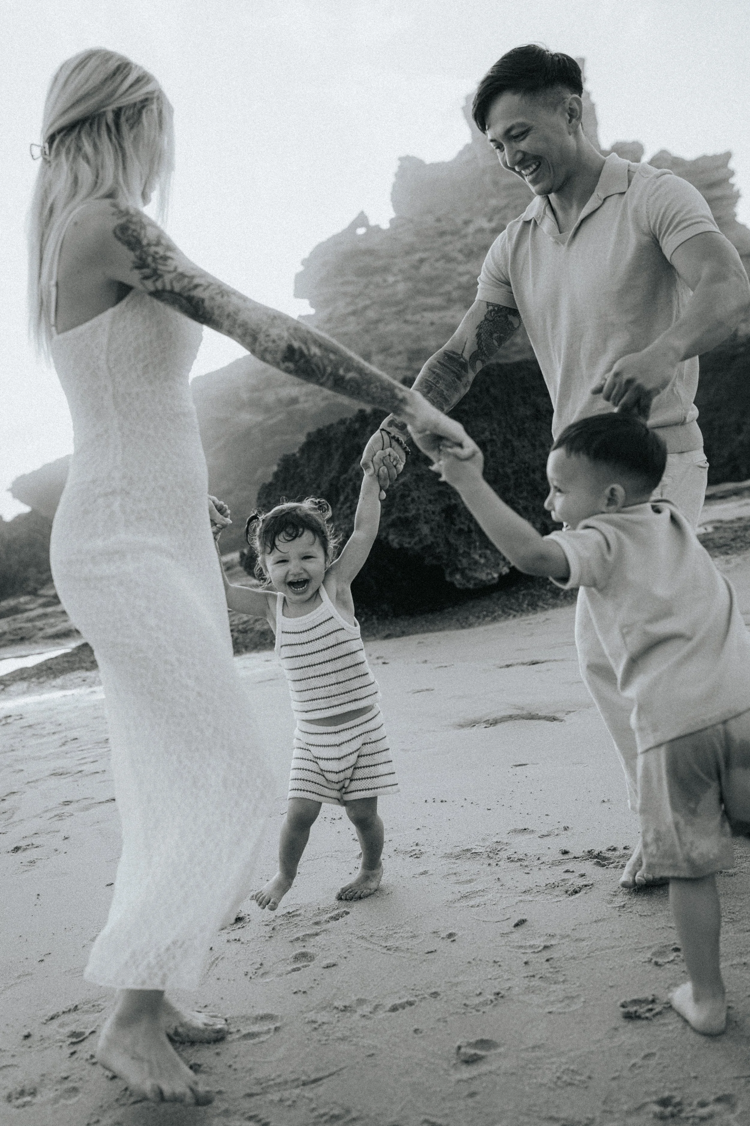 A family on the beach, holding hands and playing together, smiling and laughing, with rocky formations in the background.