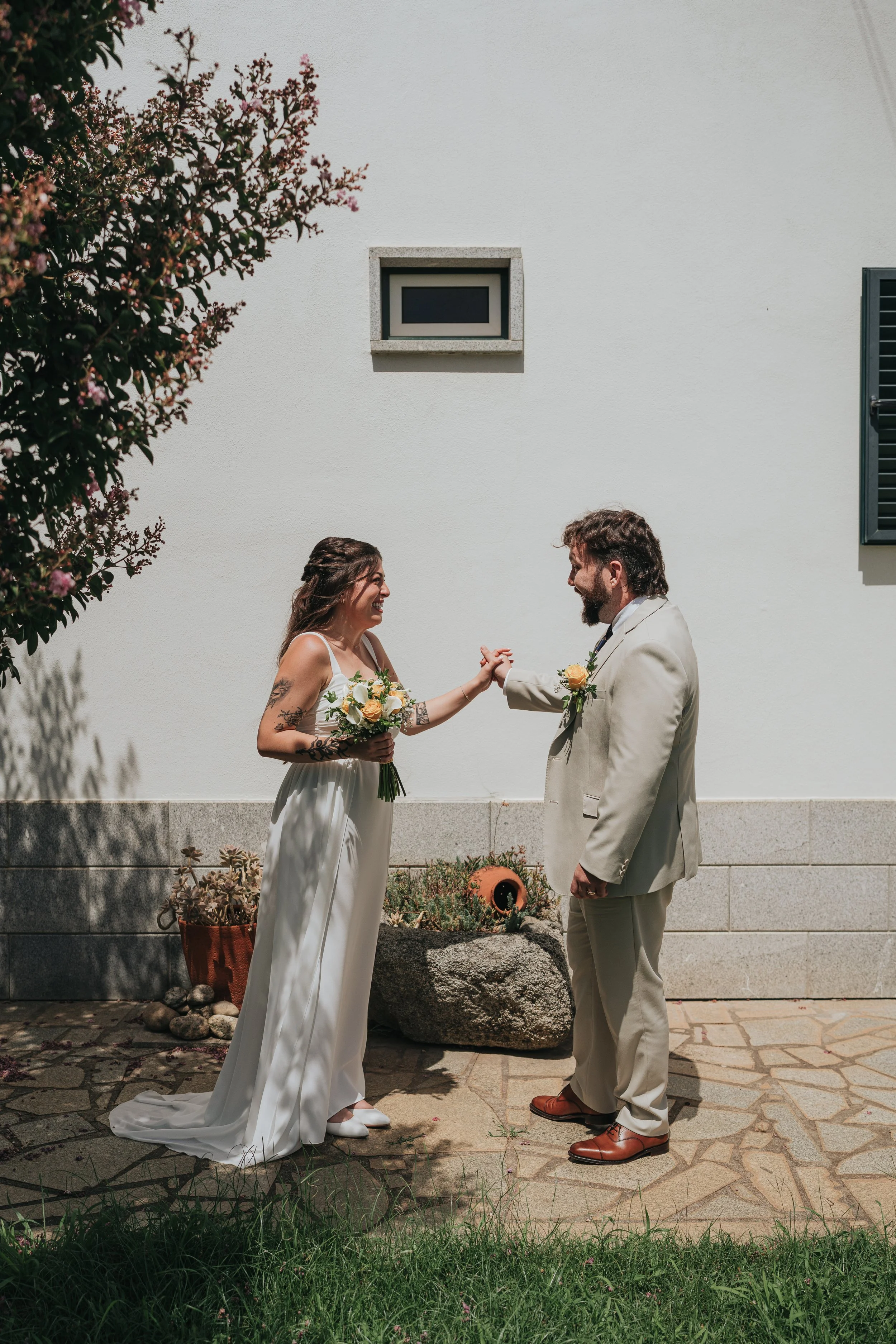 A couple dressed in wedding attire standing outdoors and holding hands, smiling at each other, against a plain white wall and some greenery.