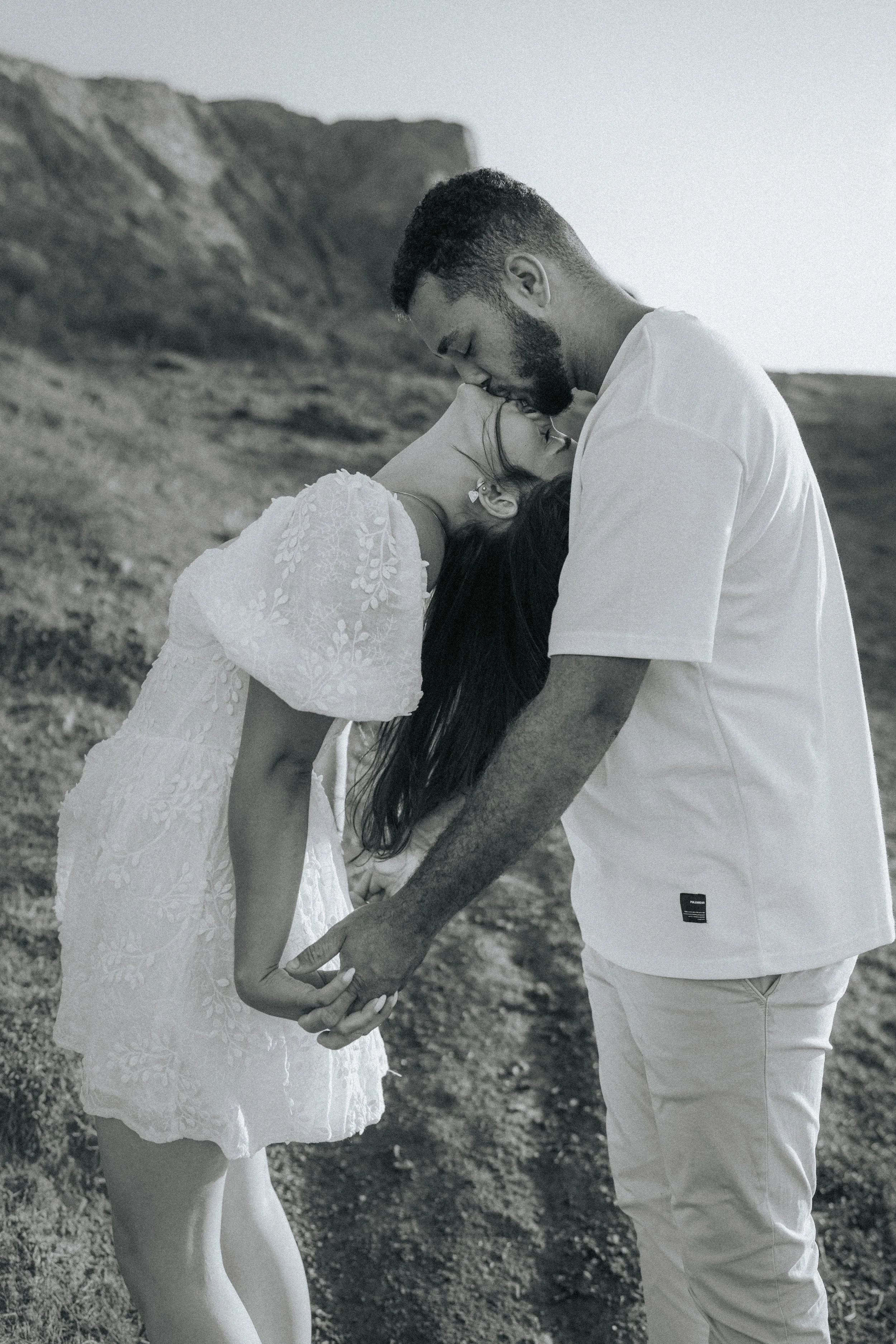 A couple holding hands, with the woman leaning her head against the man's forehead, standing outdoors on a rocky landscape. The photo is in black and white.