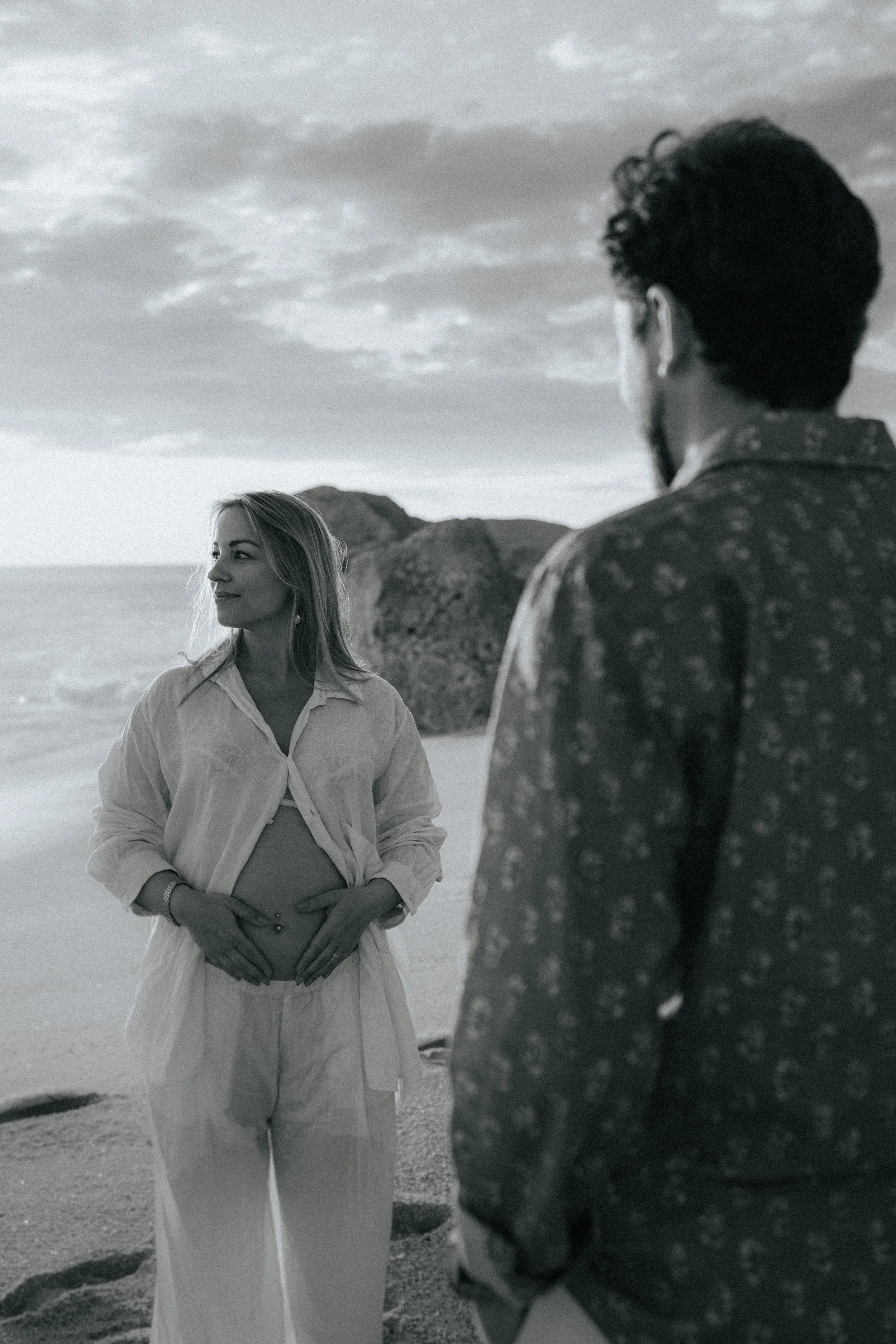 A woman with long hair wearing a light-colored top and pants, standing on a beach with her hands on her pregnant belly, looking at a man with dark, curly hair and a patterned shirt, who is turned away from the camera, with rocky landscape and cloudy 