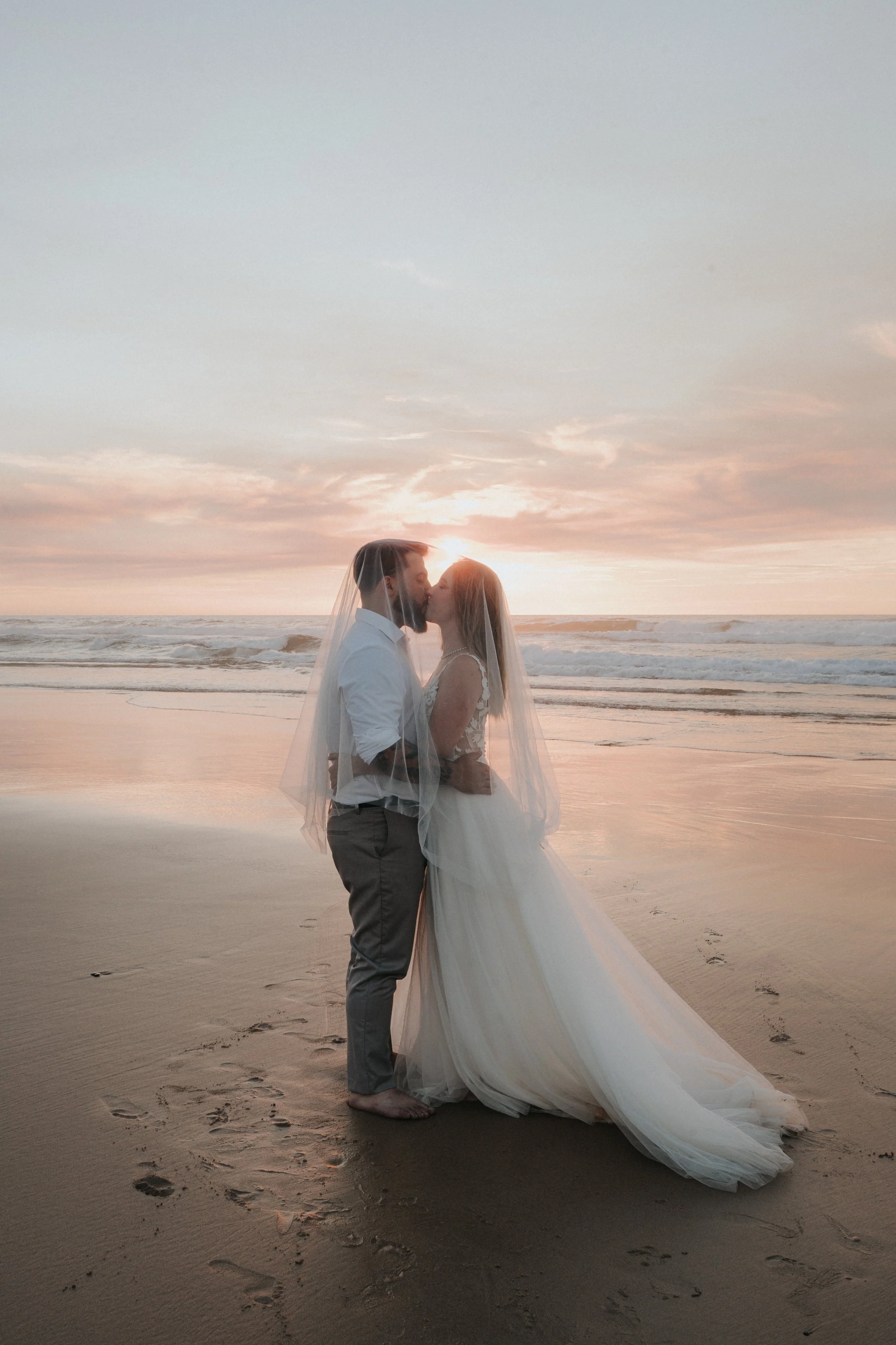 A couple in wedding attire kissing on the beach at sunset, with the bride wearing a long white dress and veil, and the groom dressed casually in a white shirt and grey trousers.