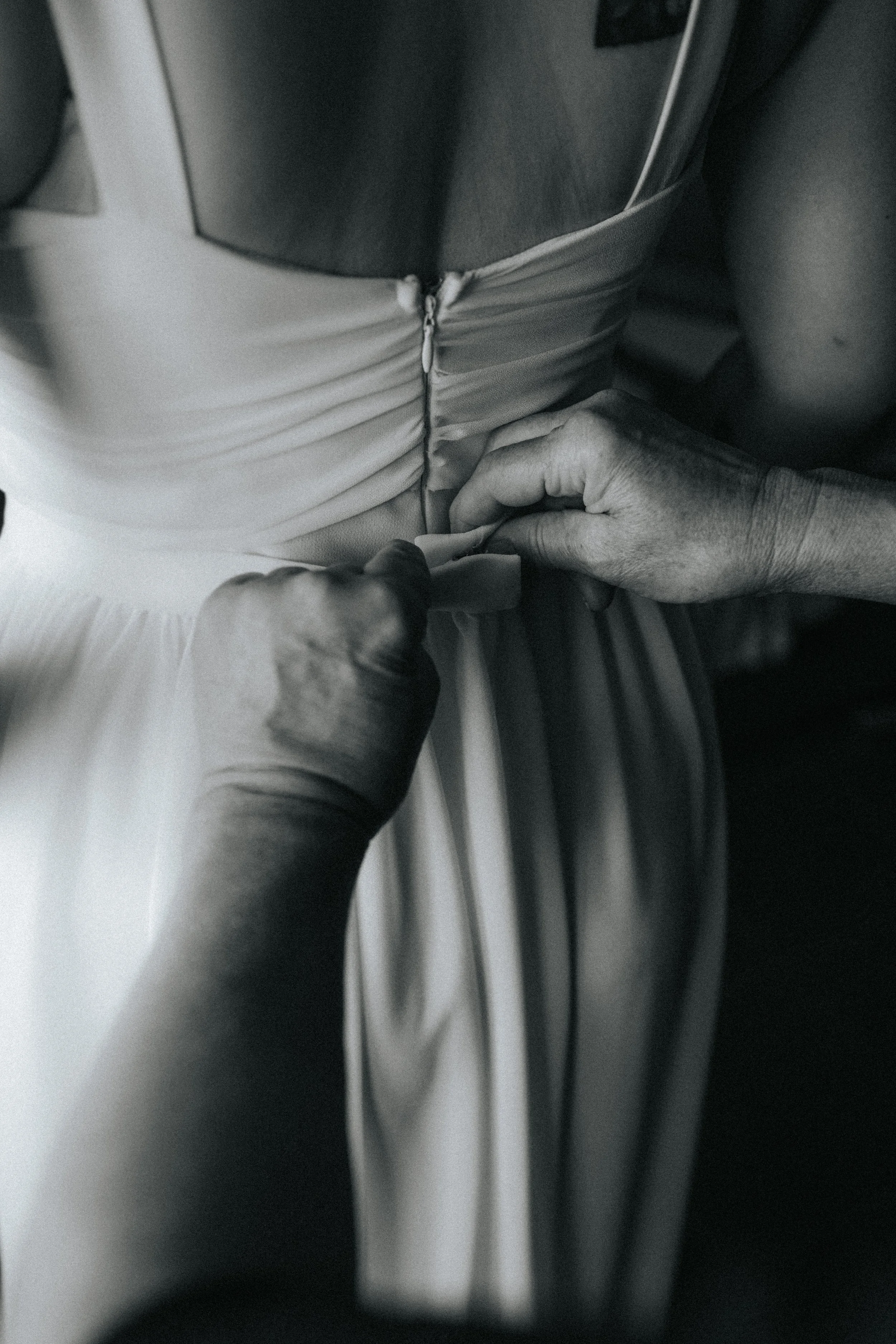 A person wearing a light-colored dress with a zipper is adjusting the zipper with their hands, while sitting. The image is in black and white.