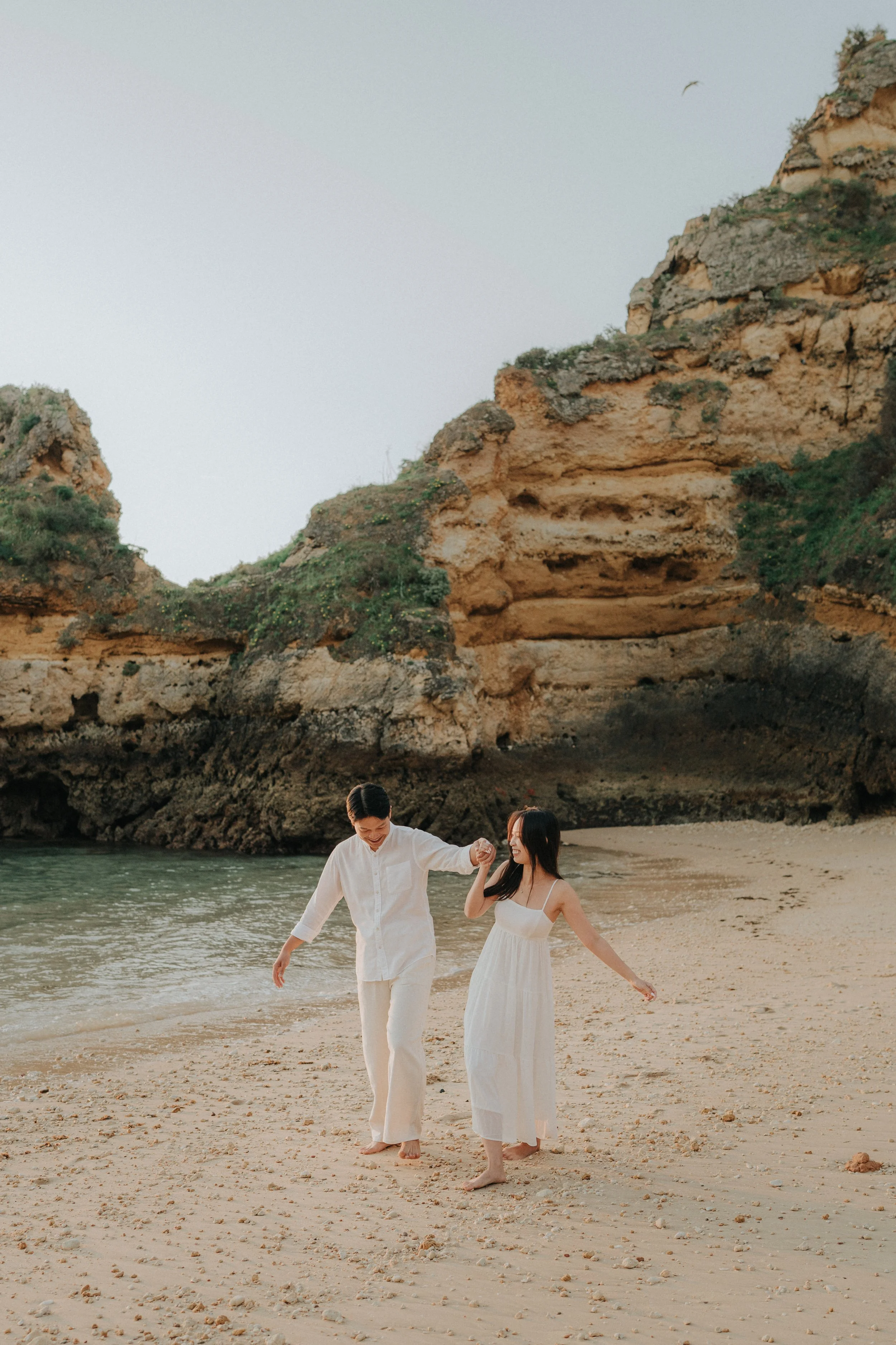 A young couple in white clothing holding hands and walking along the beach, with rocky cliffs and a seagull flying overhead in the background.