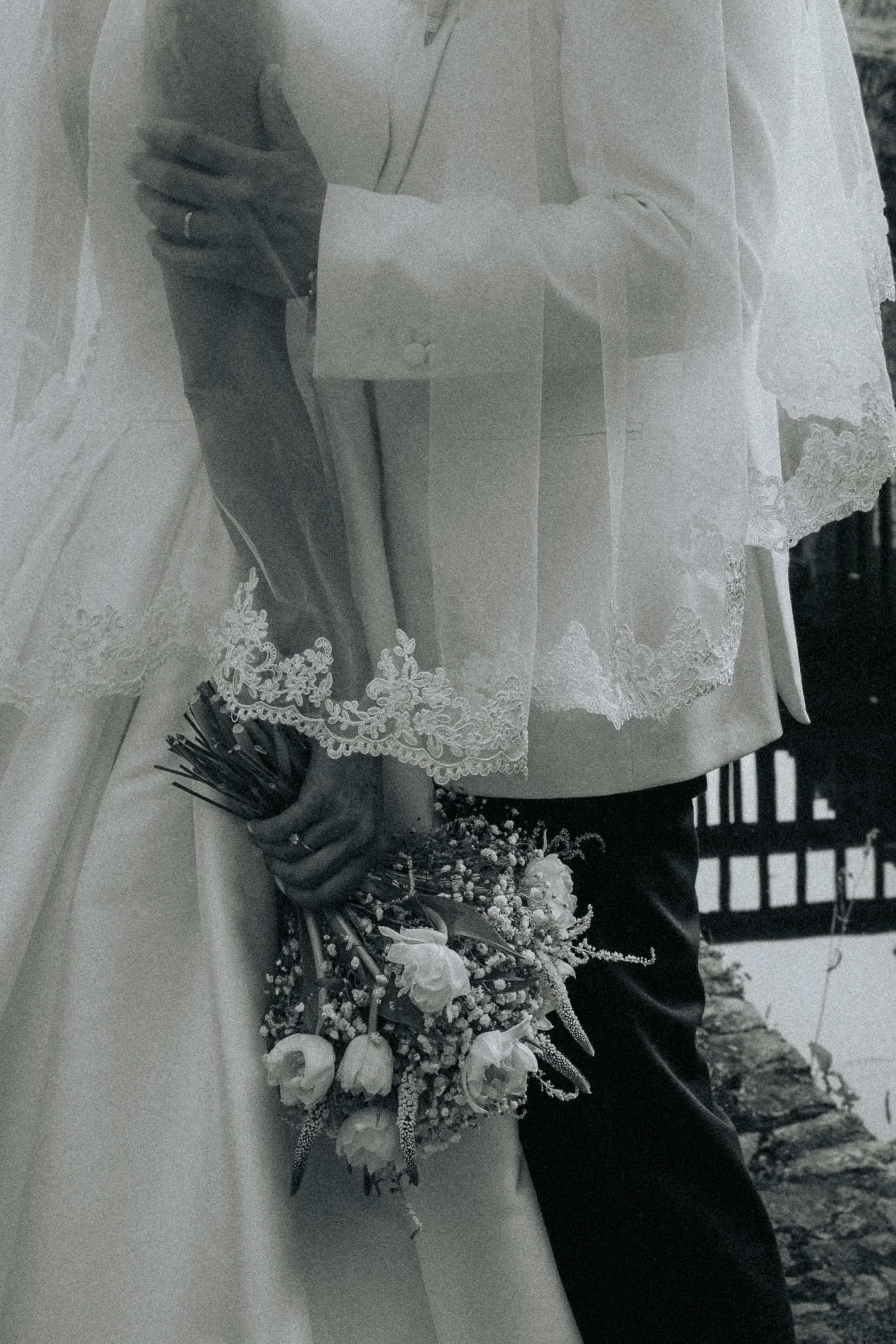 A bride in a white lace dress holding a bouquet of flowers, standing close to a man in dark clothing, possibly during a wedding ceremony.