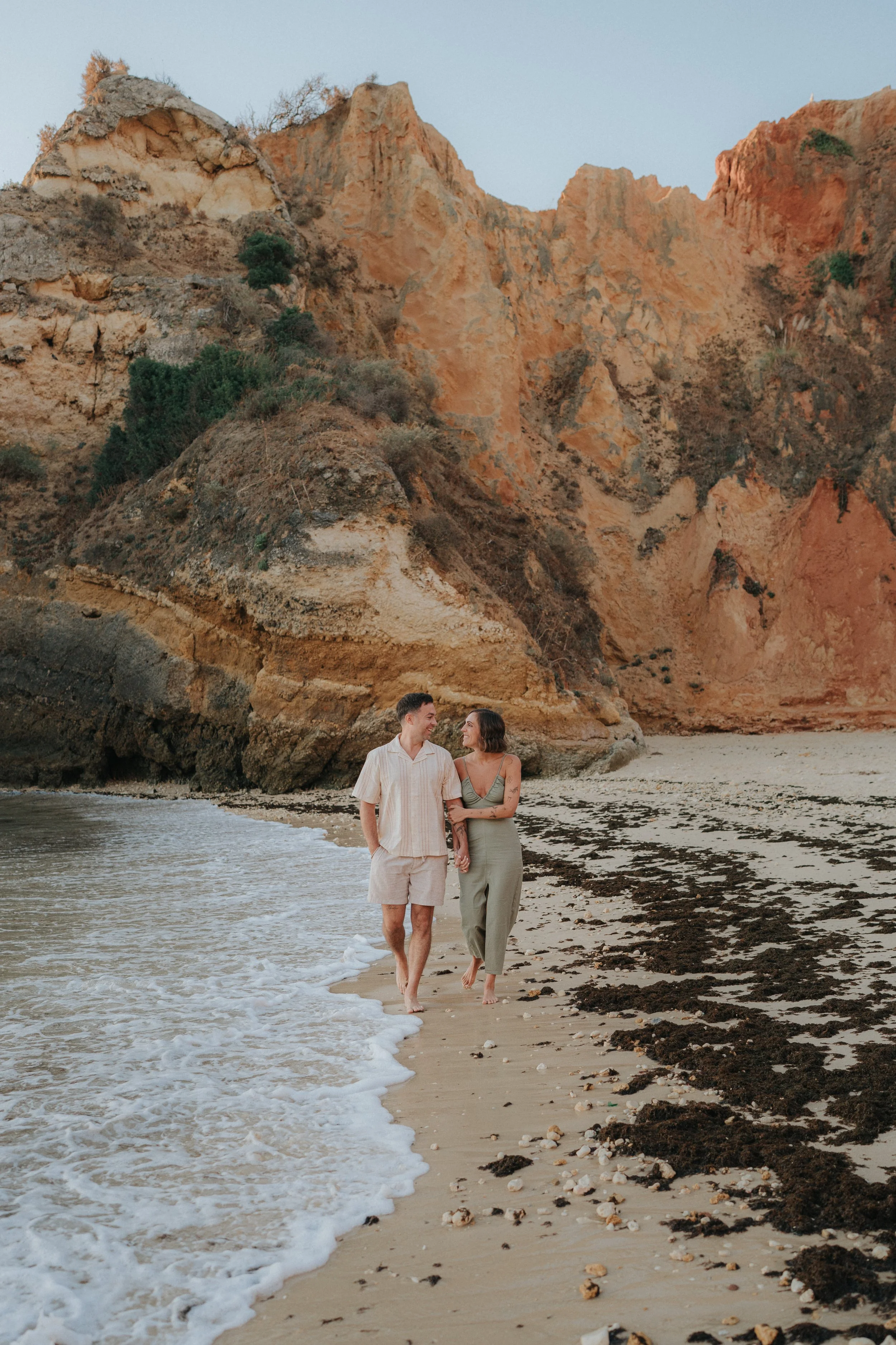 A couple walking along a sandy beach with dark seaweed and shells, with tall orange and beige rocky cliffs behind them, under a clear sky.