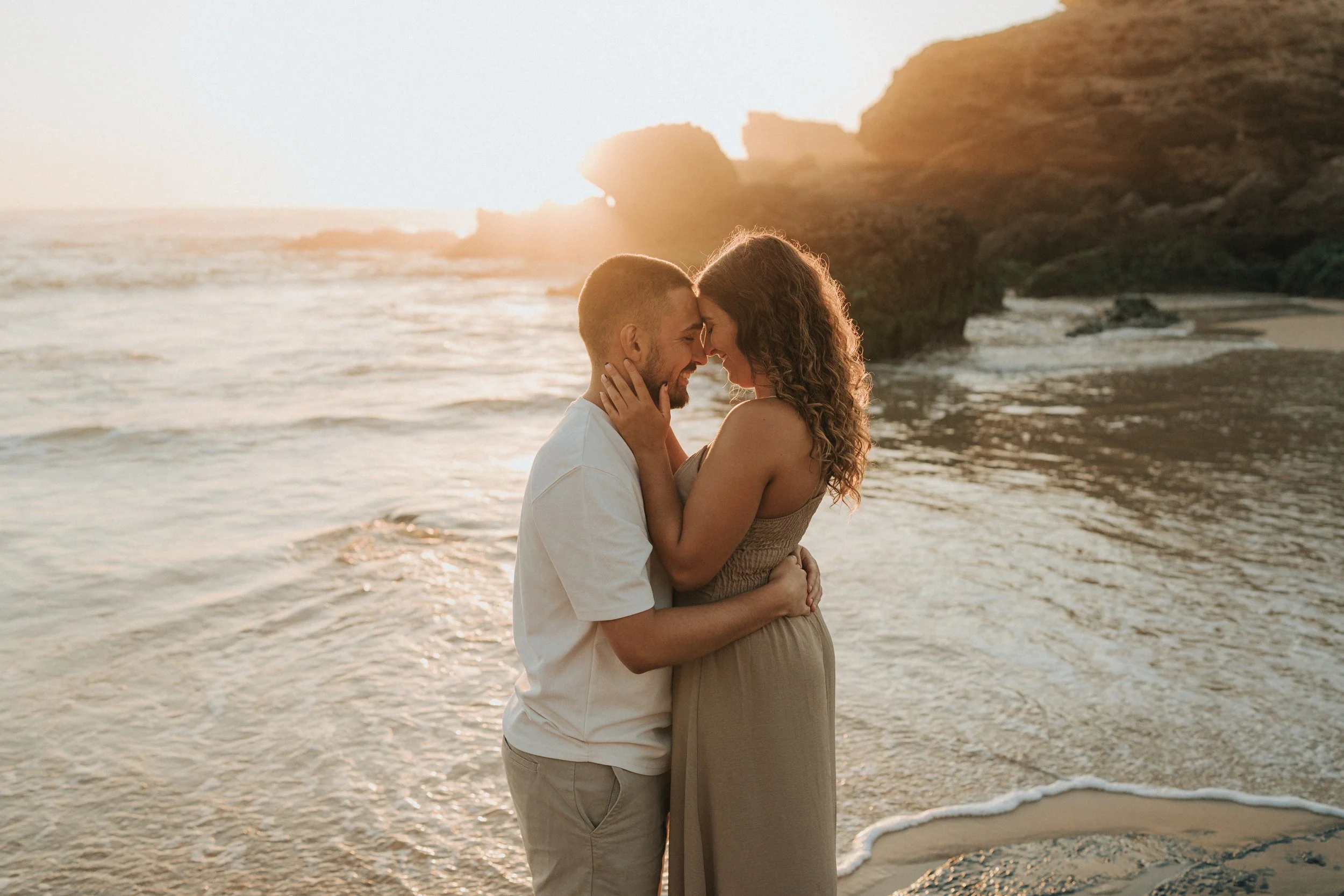 A couple on the beach at sunset, embracing and touching foreheads with their eyes closed.