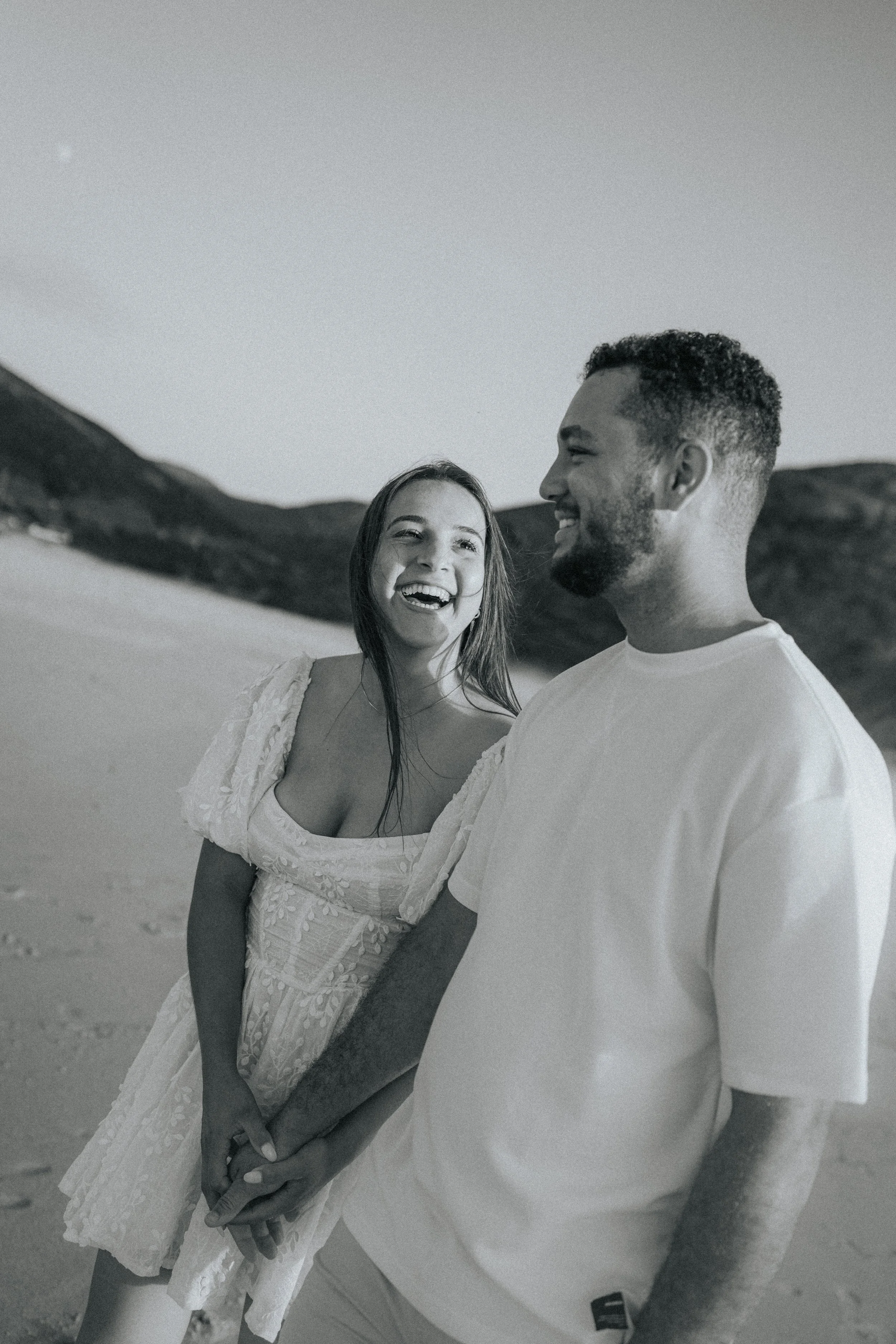 A smiling woman and a man holding hands on a beach, facing each other, with mountains in the background.