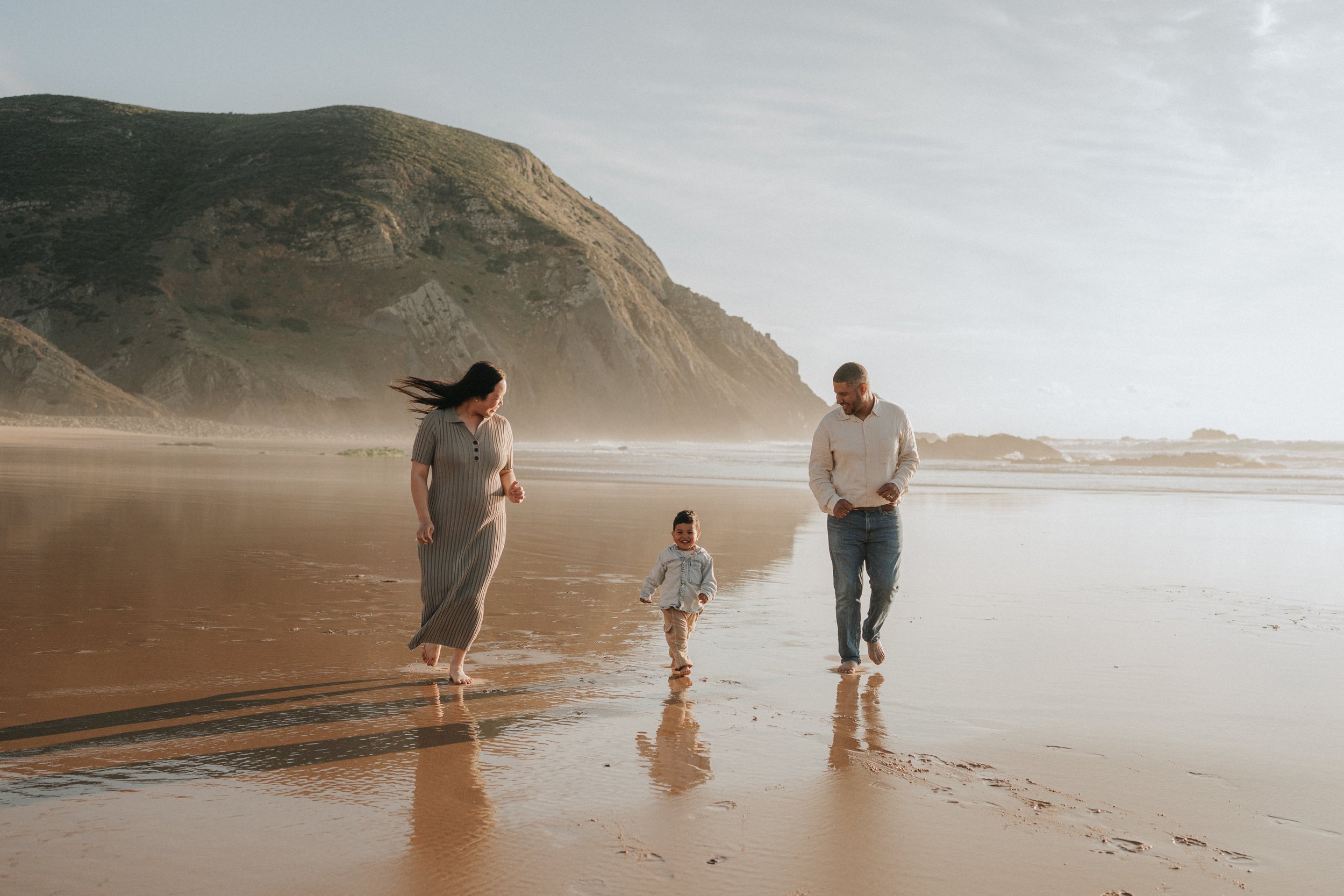 A family of four walking barefoot on a beach near the water with hills in the background. The woman is wearing a long striped dress, and the man is dressed in a light shirt and jeans. Two children, a boy and a girl, walk nearby, smiling and enjoying 