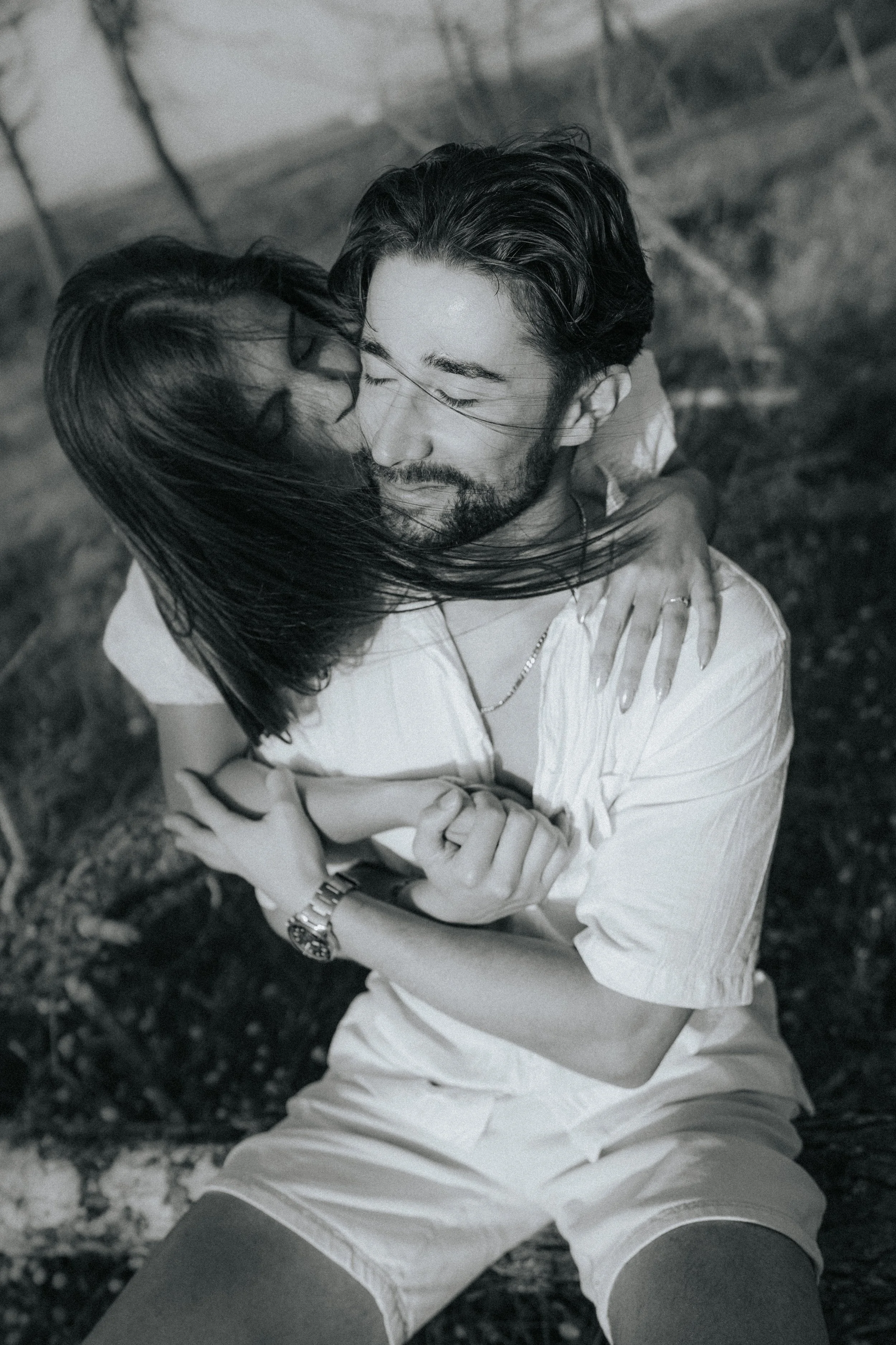 A black and white photo of a man and woman hugging tightly. The man has long, dark hair and a beard, and is wearing a light-colored shirt. The woman has long hair, glasses, and is wearing a dark top. They are outdoors, possibly in a park or garden, w
