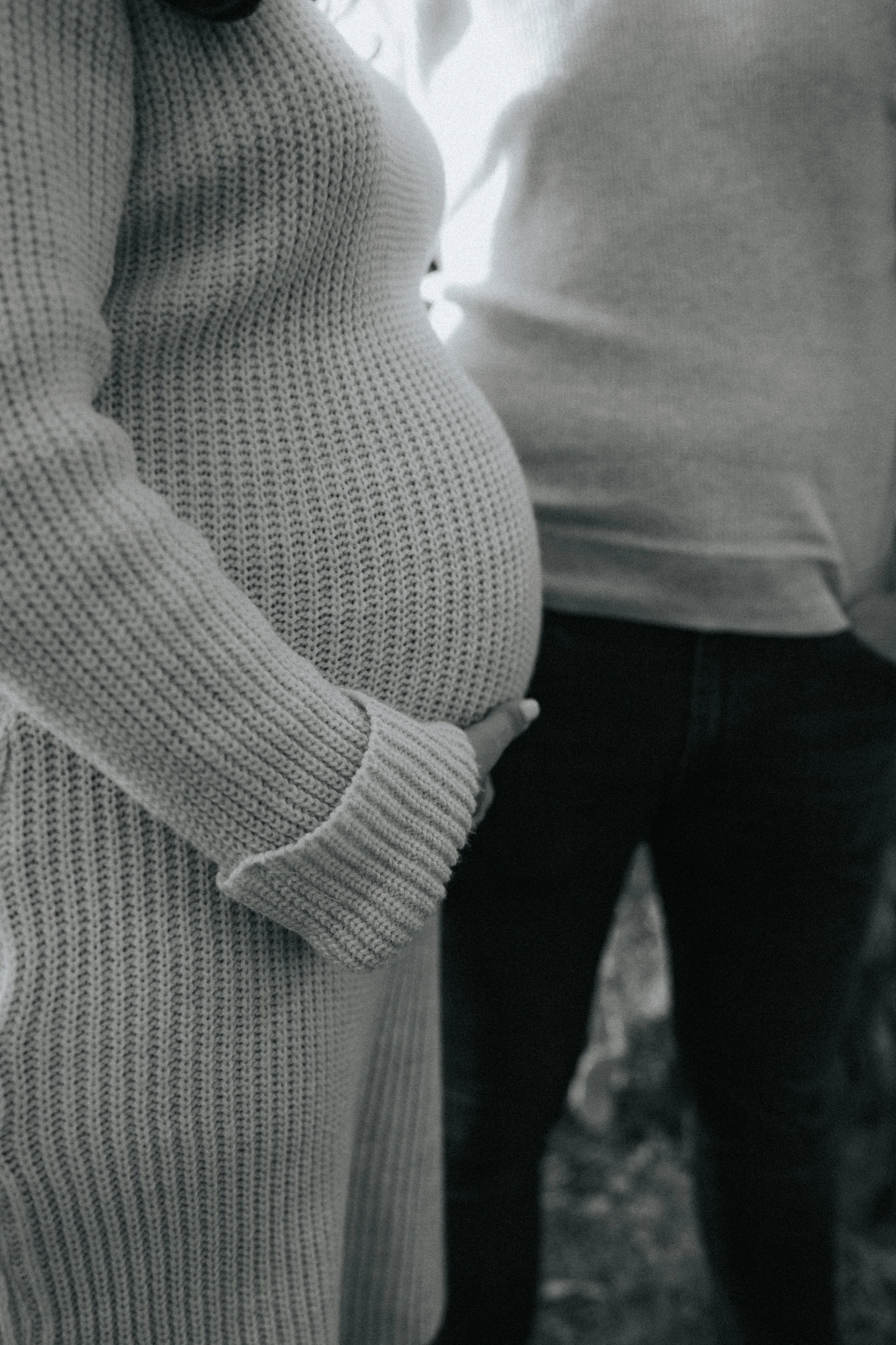 A close-up of a pregnant woman wearing a knitted sweater, cradling her belly, with another person standing close by in a casual setting.