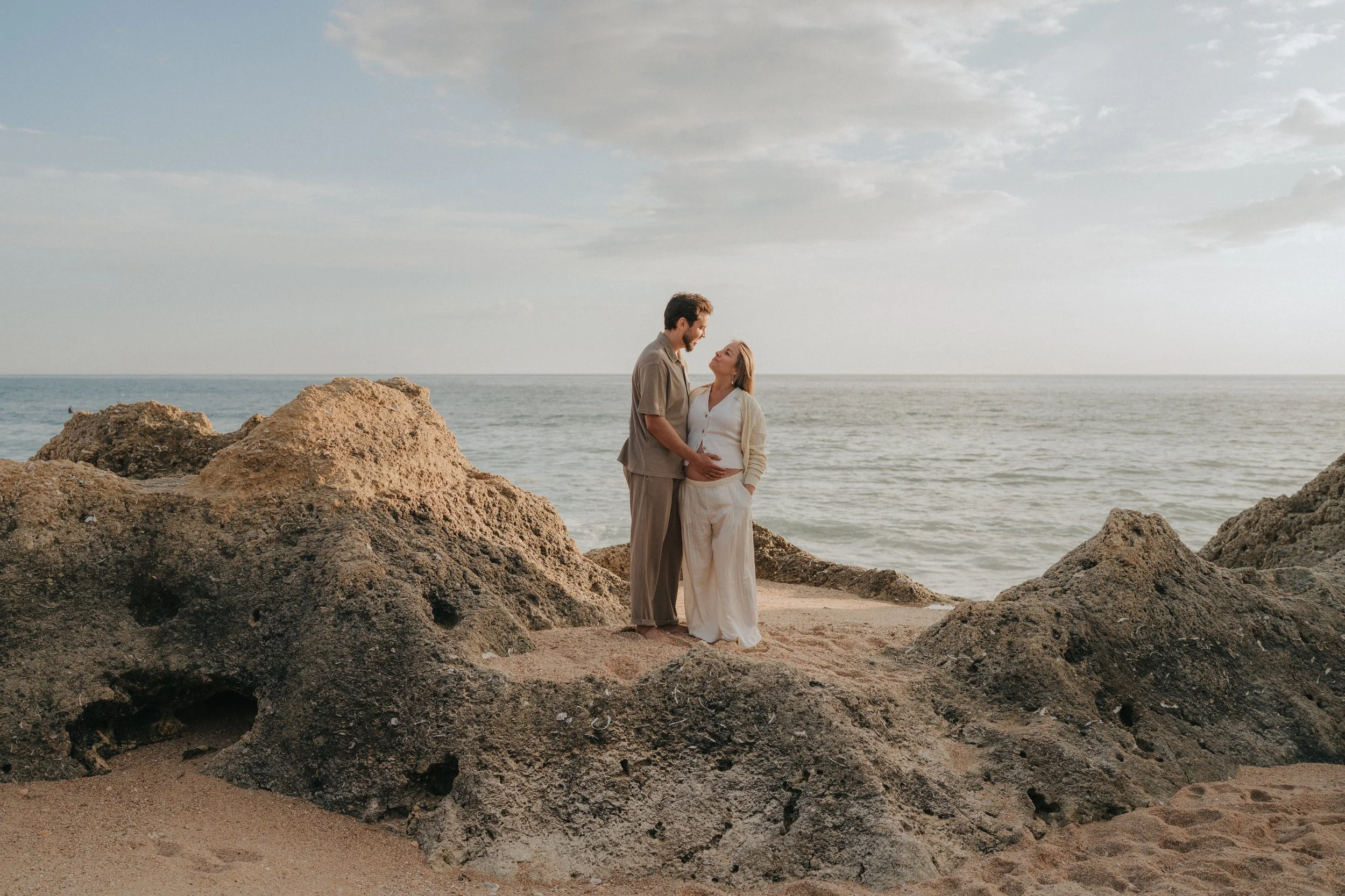 A man and woman stand close together on a beach, gazing into each other's eyes, with ocean and cloudy sky in the background.