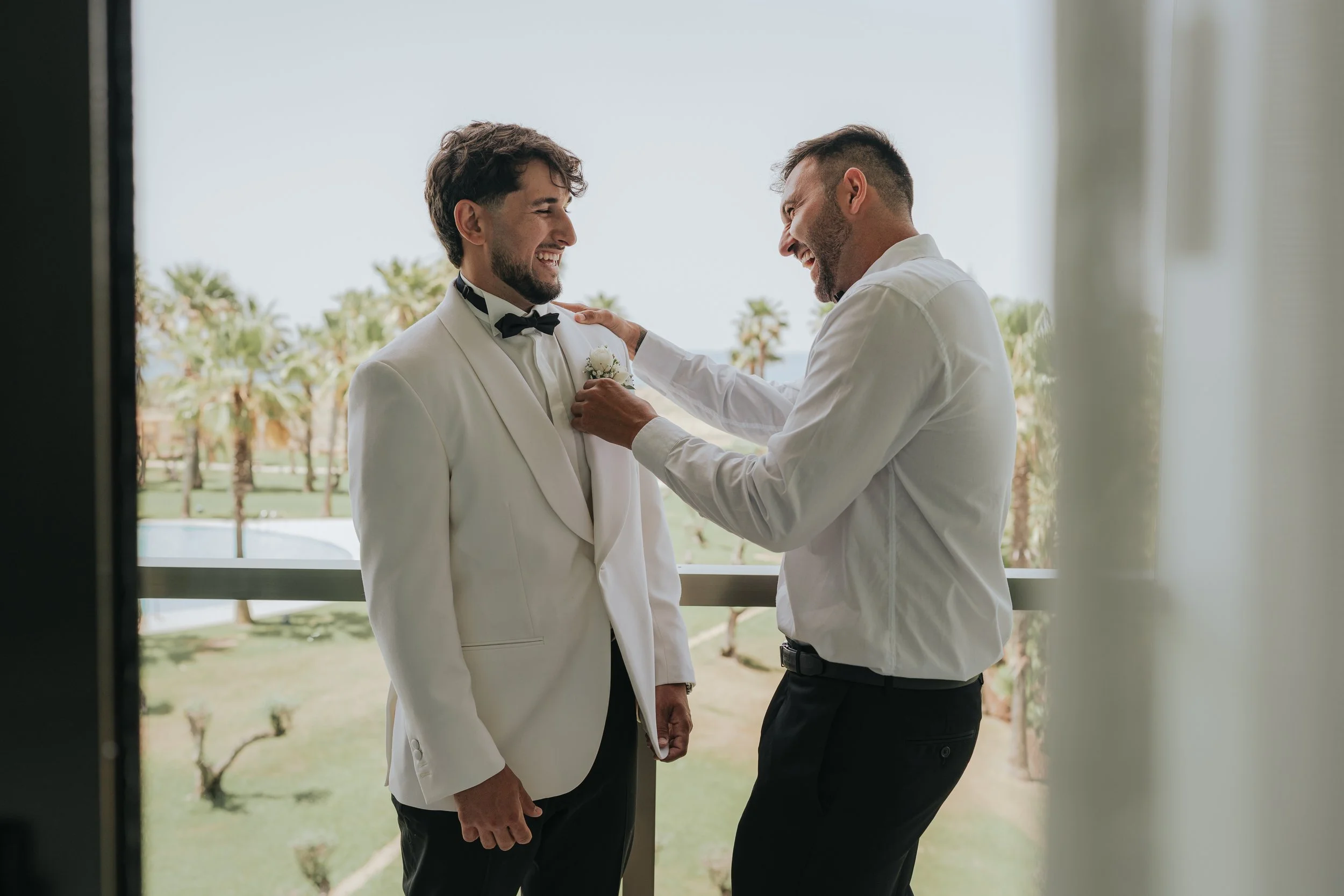 Two men dressed in formal attire, including white shirts and black ties, are smiling and interacting positively. One appears to be adjusting the other's jacket or boutonniere, with a background of palm trees and a bright, sunny sky.