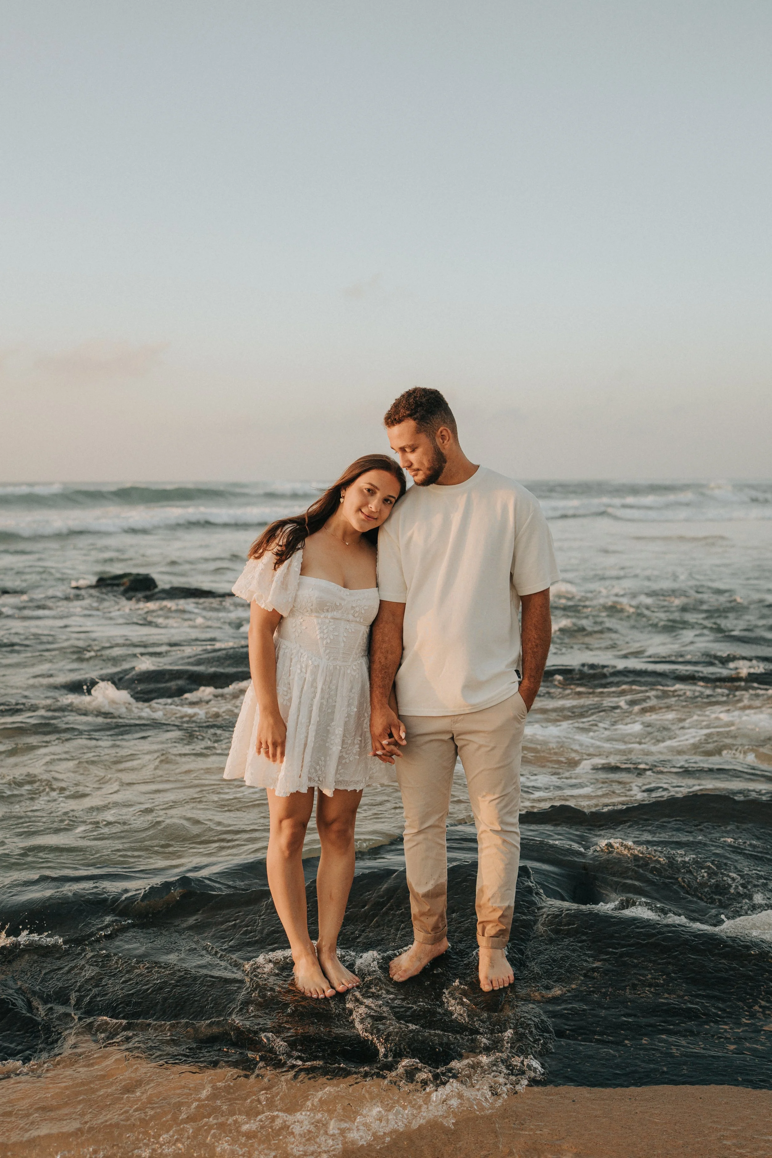 A young couple holding hands and standing in the ocean at sunset, with the woman in a white dress and the man in a white t-shirt and beige pants, both barefoot.