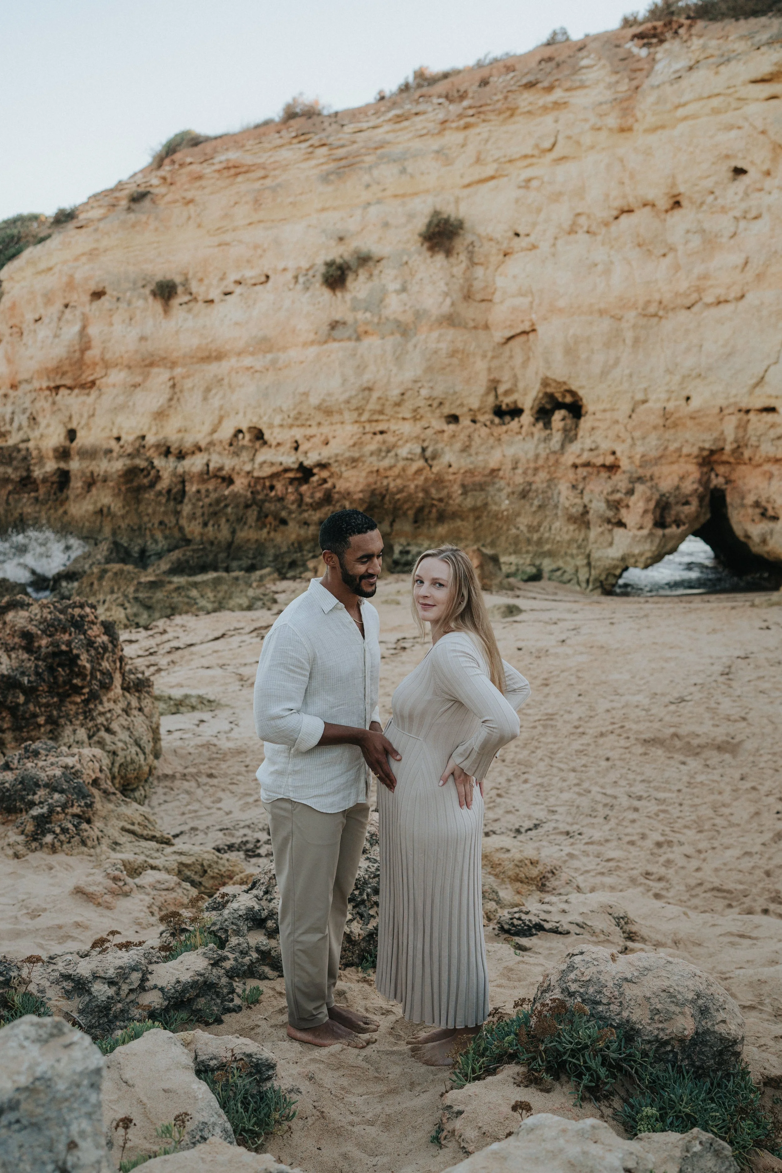 A couple standing on a sandy beach with rocky formations and a large sandstone cliff in the background. The man has dark skin, dark hair, and a beard, wearing a light-colored long-sleeve shirt and beige pants. The woman has light skin, blonde hair, a