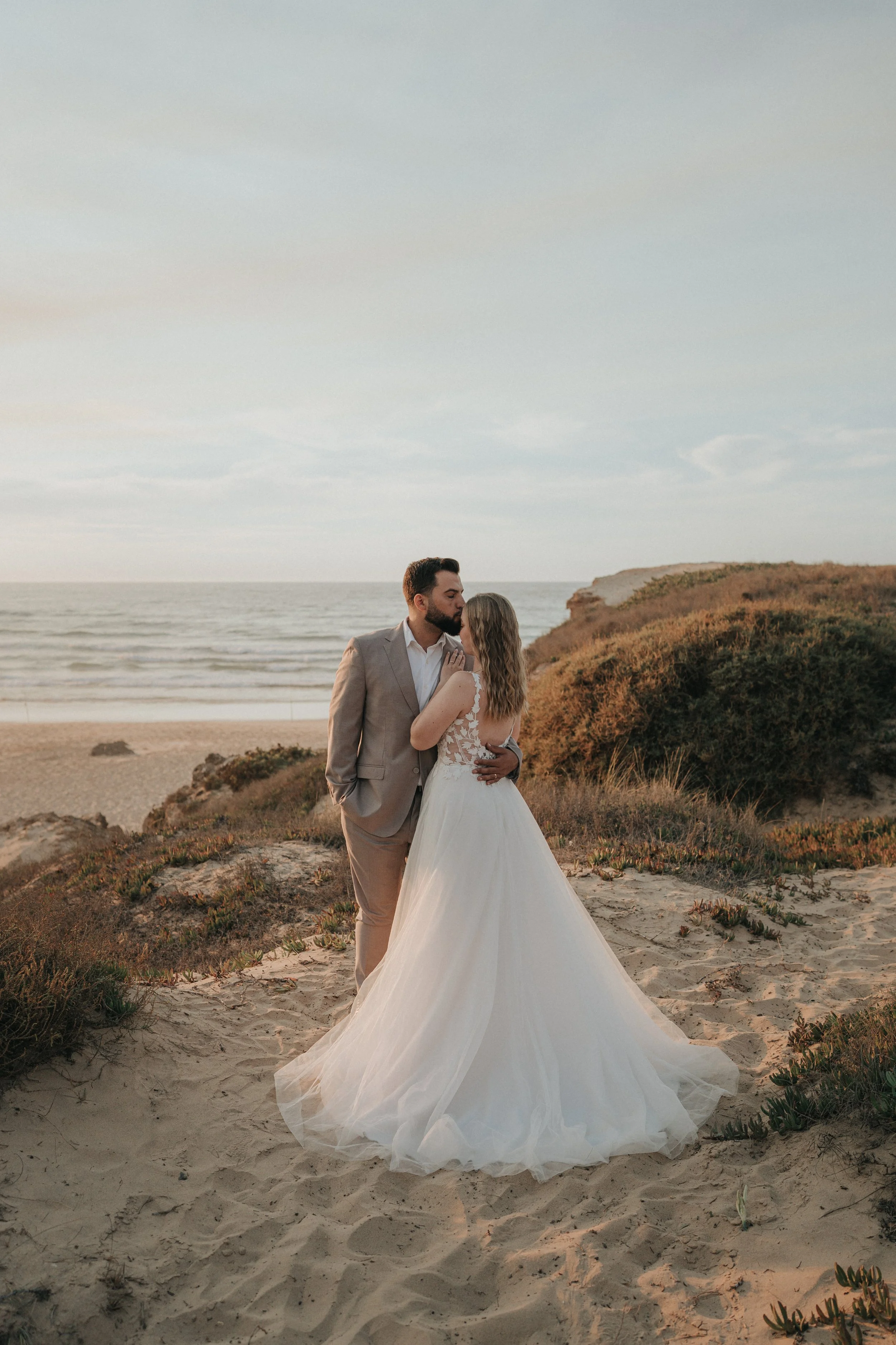 A couple in wedding attire standing on a sandy beach with ocean waves and cliffs in the background during sunset.
