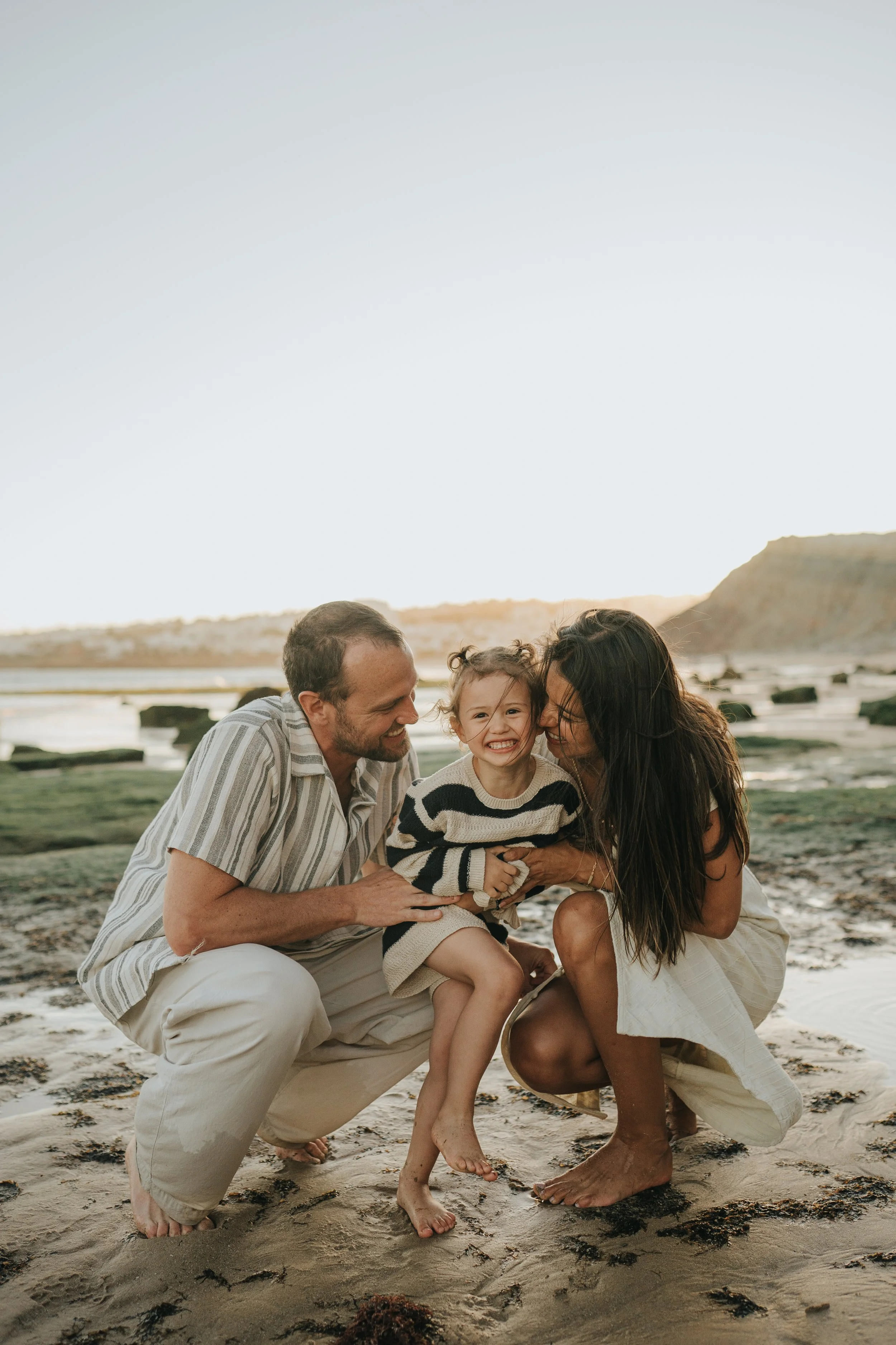 A happy family of three, including a man, woman, and young girl, kneeling and sitting on the beach at sunset, smiling and hugging each other.