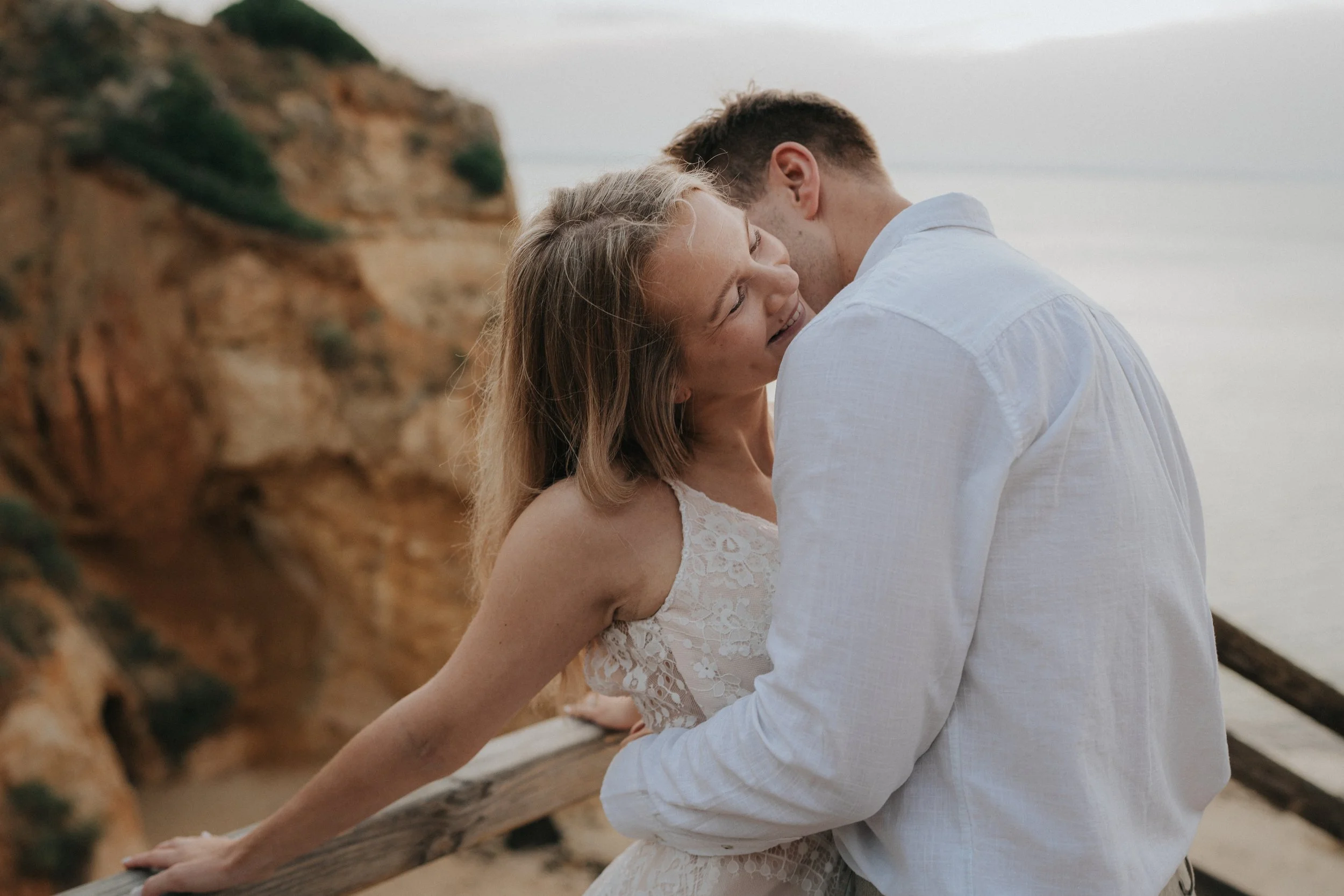 A couple embraces on a beach, with a rocky cliff in the background, during sunset. The woman wears a lace white dress, and the man wears a white shirt.
