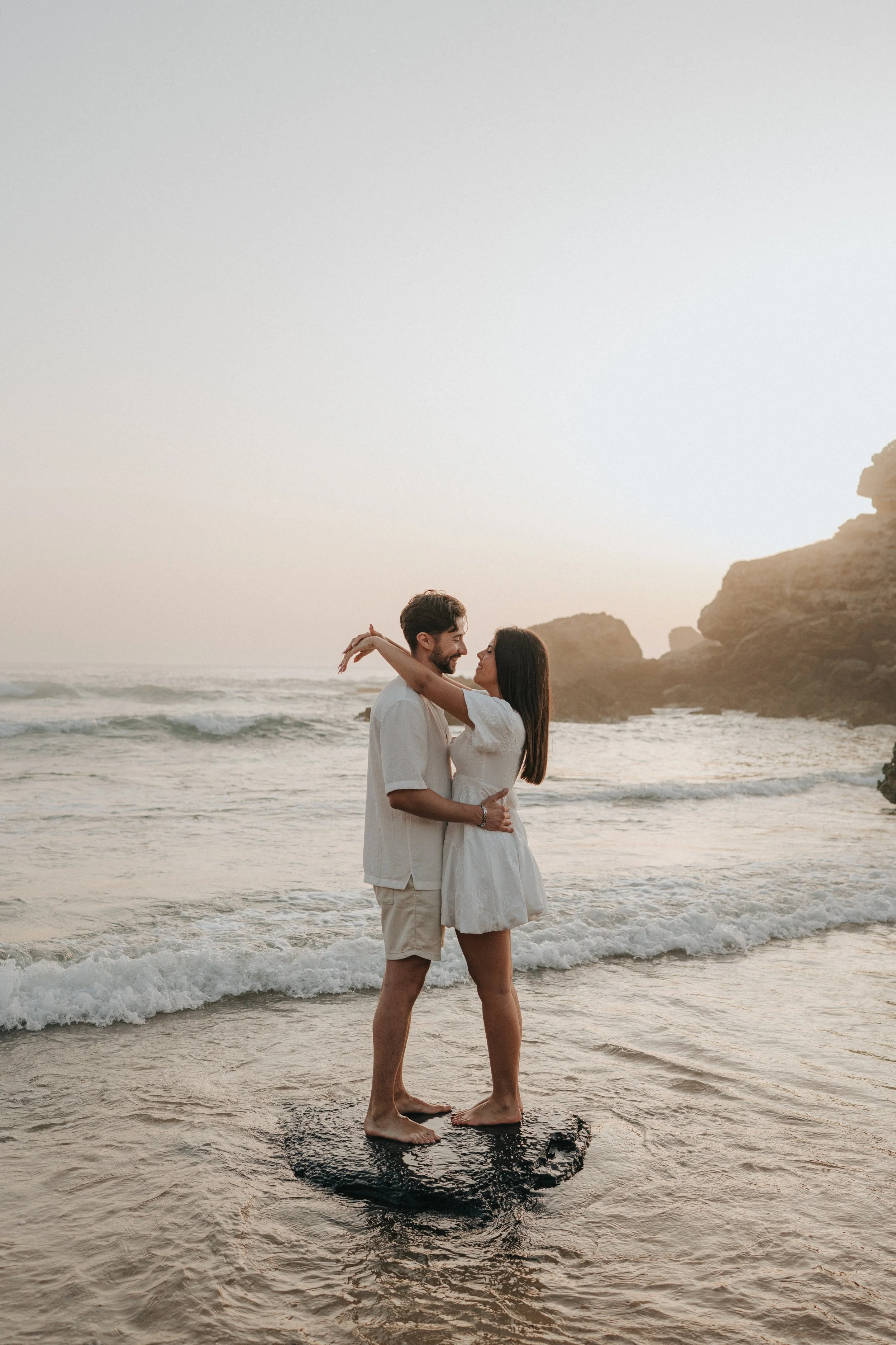 A couple embracing and looking at each other on the beach, standing on a surfboard with ocean waves and rocky cliffs in the background during sunset.