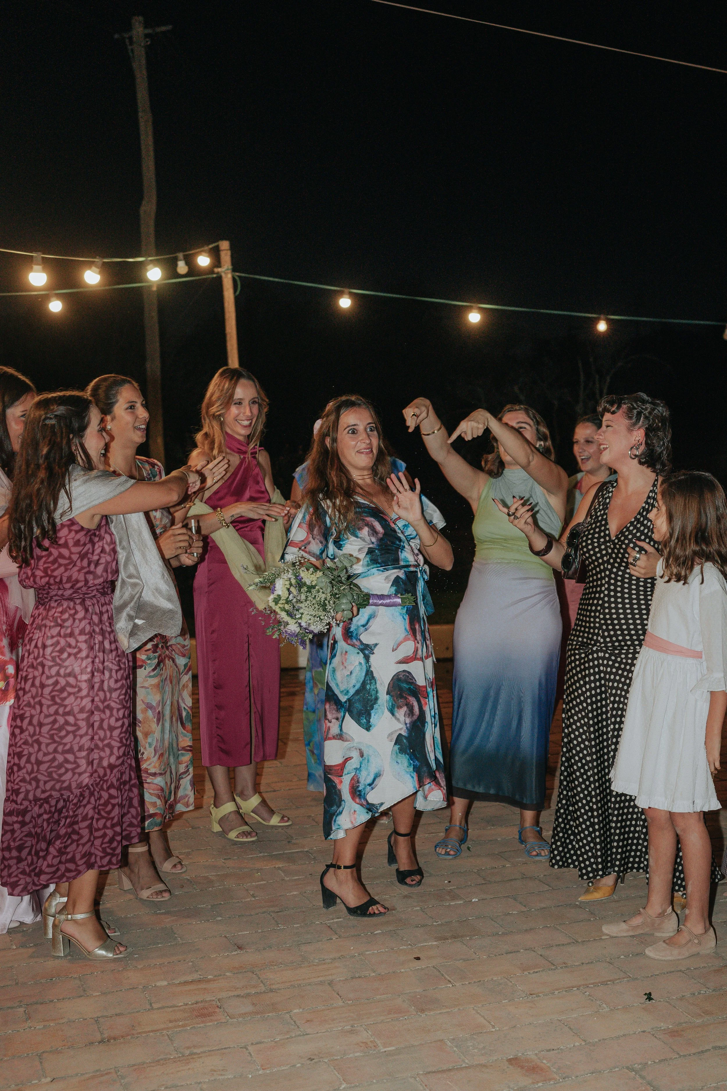 A group of women dressed in colorful, fashionable dresses are gathered outdoors at night, smiling and socializing under string lights. One woman is holding a bouquet of flowers.