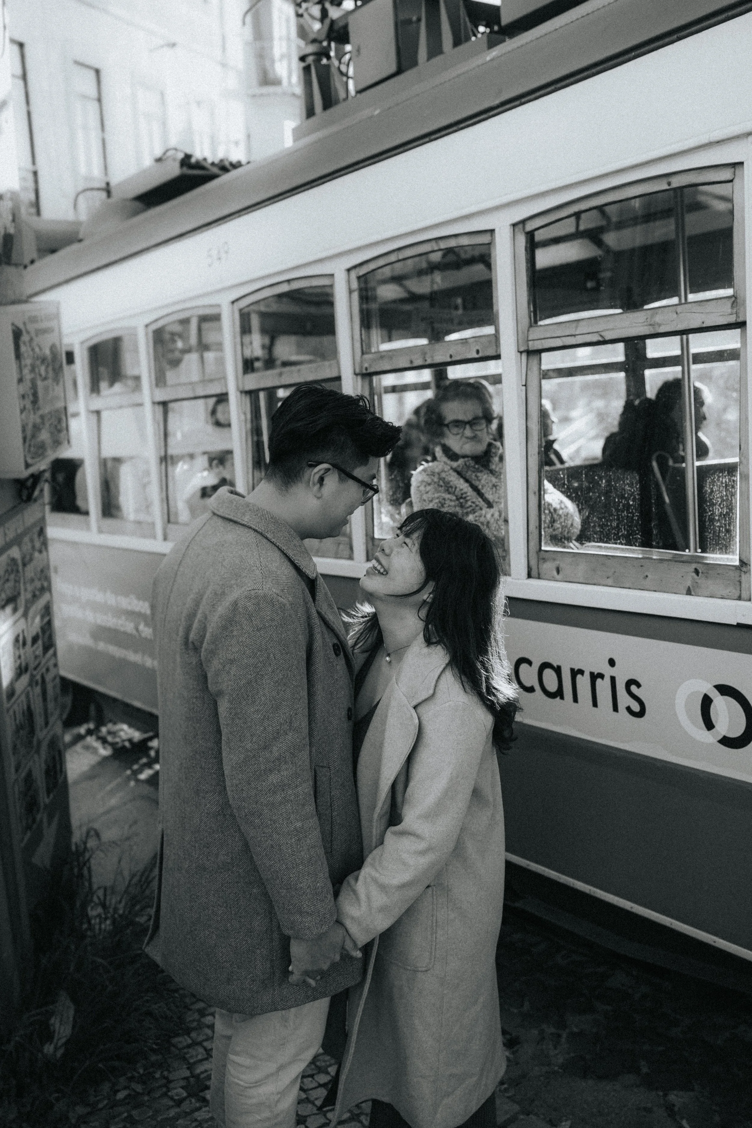 A black and white photo of a couple holding hands and embracing each other. The man is wearing glasses and a coat, and the woman is smiling and looking up at him. They are standing outdoors near a streetcar or tram with visible windows and signage.