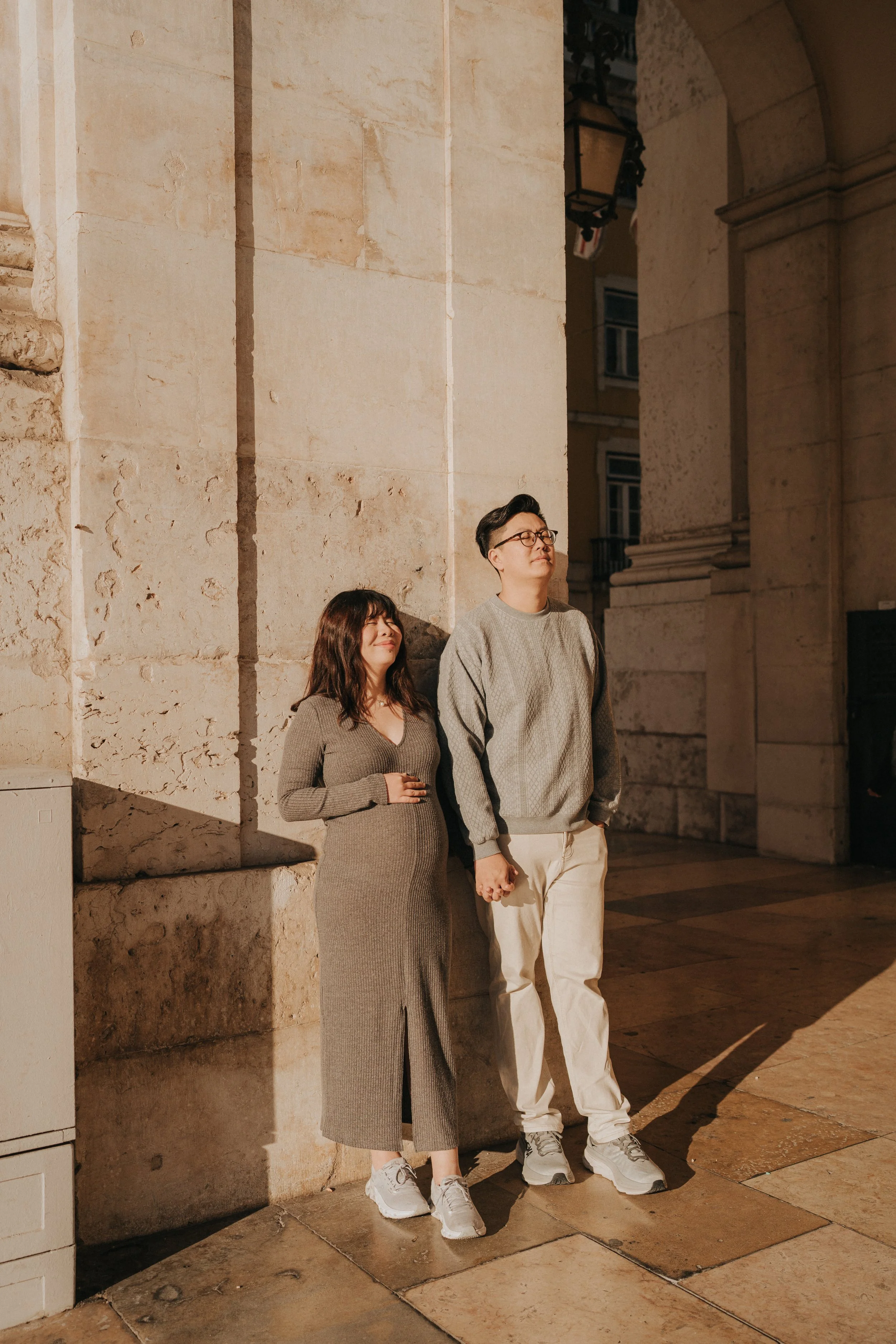 A man and woman holding hands and standing against a stone wall outside at night, with the woman smiling and the man with eyes closed, in a warm moment.