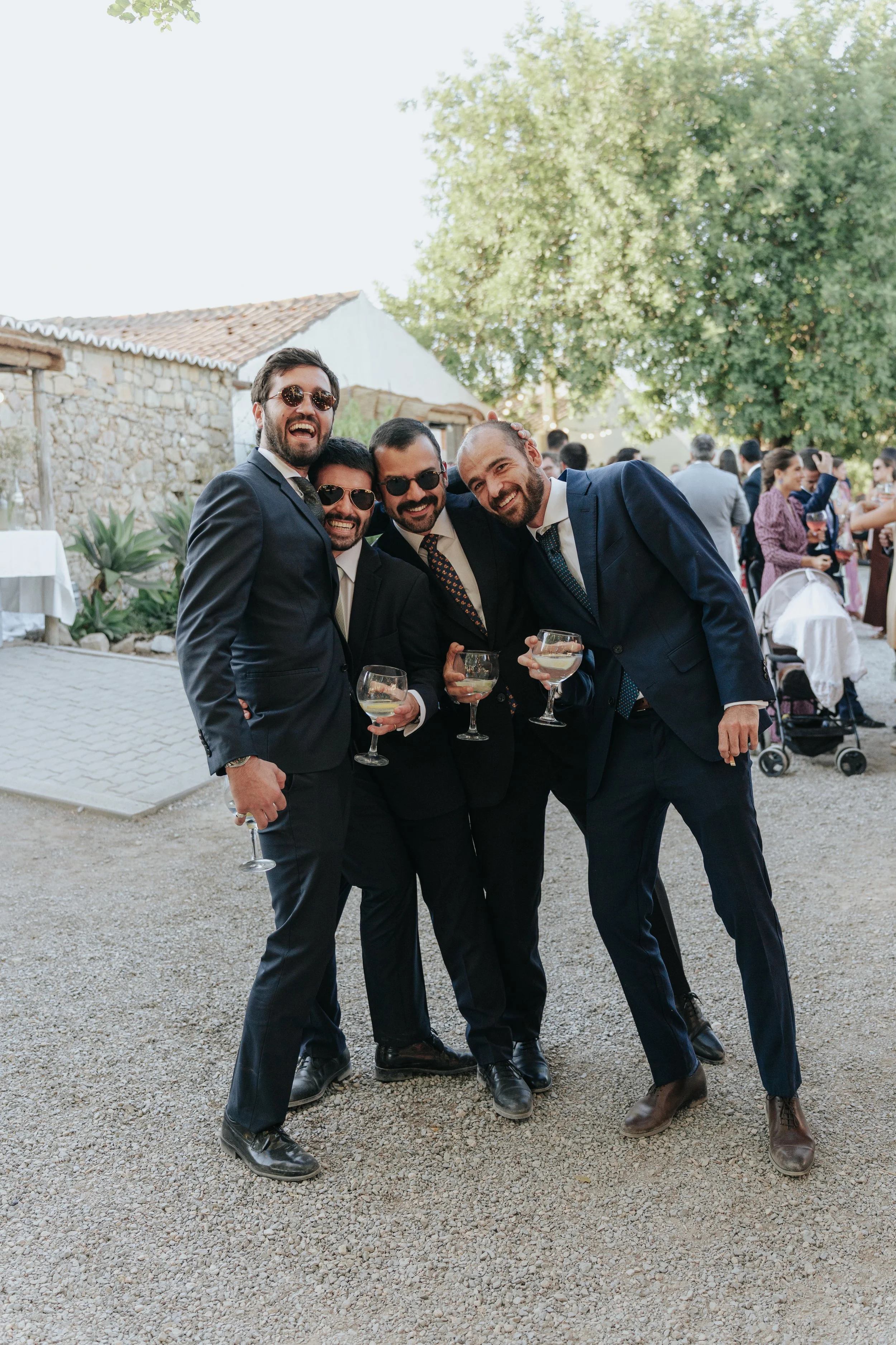 A group of four men in suits and sunglasses celebrating at an outdoor event, holding glasses of drinks, with a crowd of people and greenery in the background.