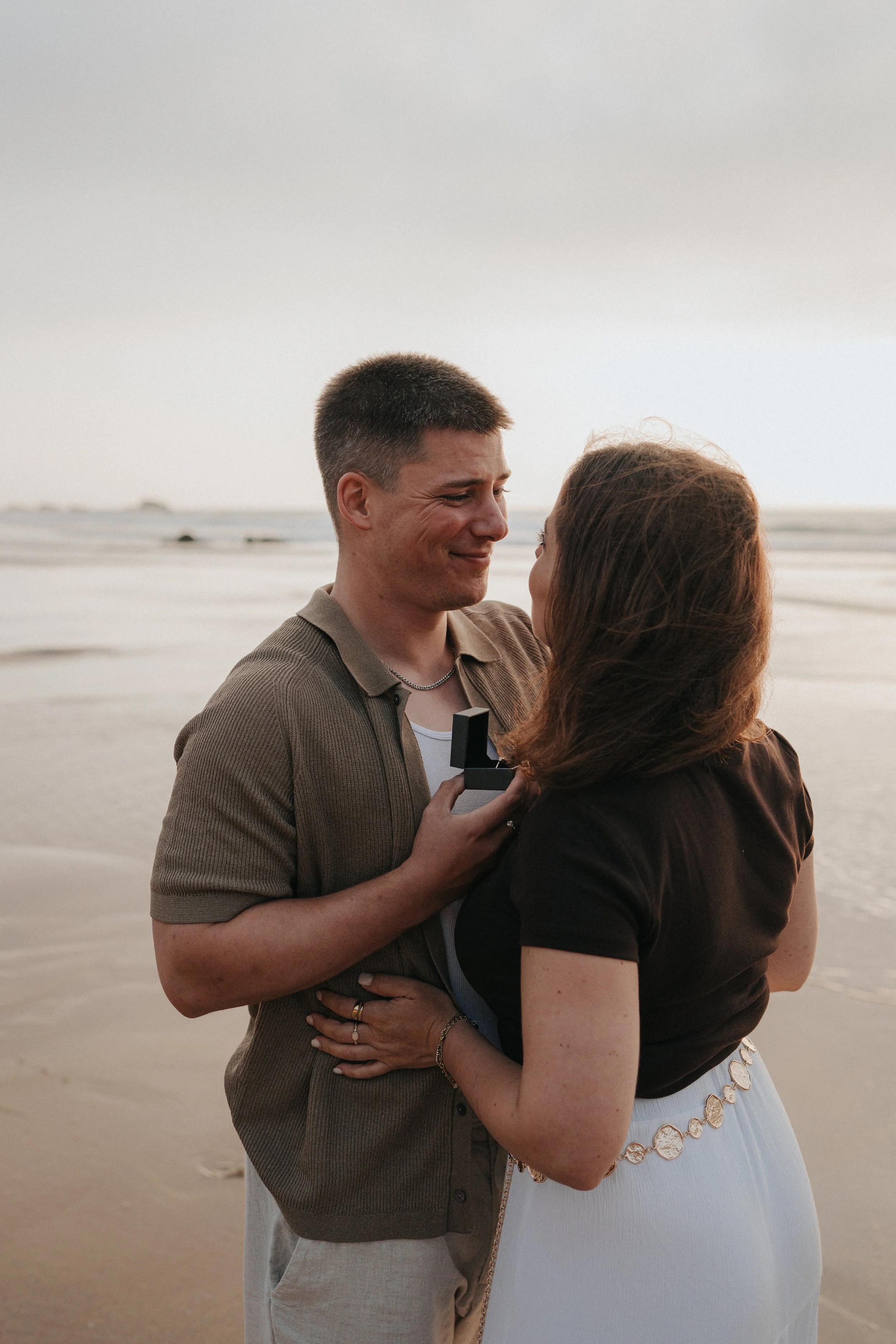 A couple stands on a beach during sunset, gazing into each other's eyes, holding each other tenderly. The man is in a brown shirt, and the woman is in a black top and white skirt with a circular pattern, both with light skin. The background shows the