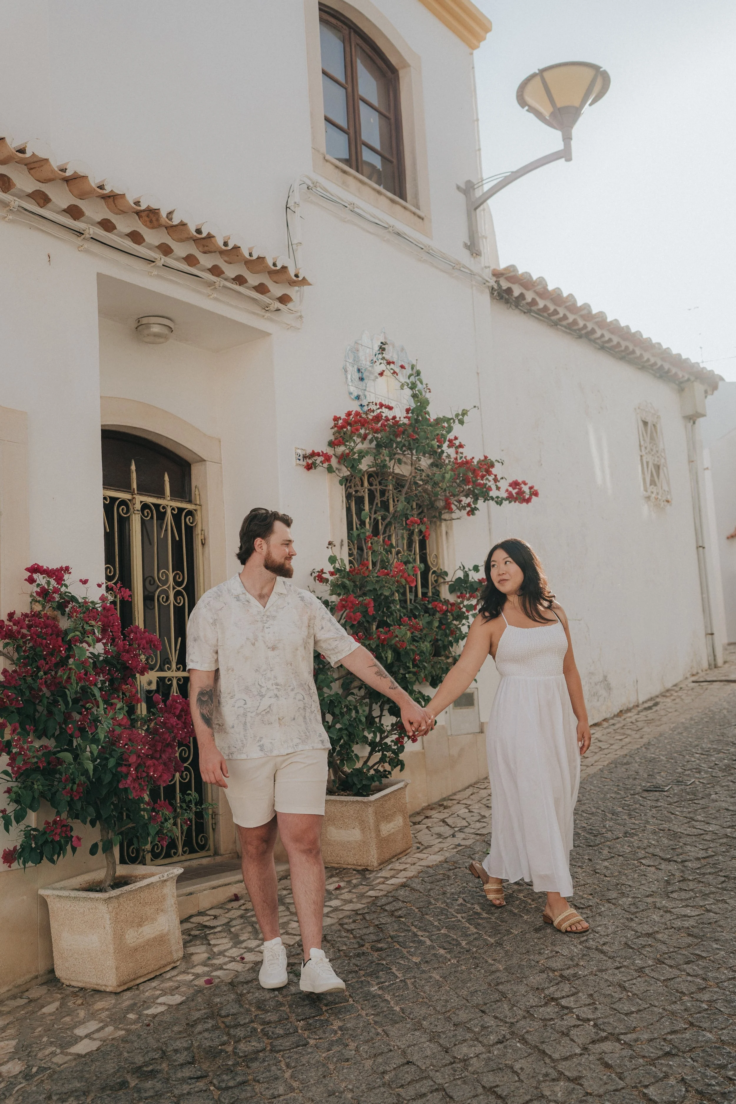 A couple holding hands walking on cobblestone street outside a white building with flower pots and greenery, under a street lamp.
