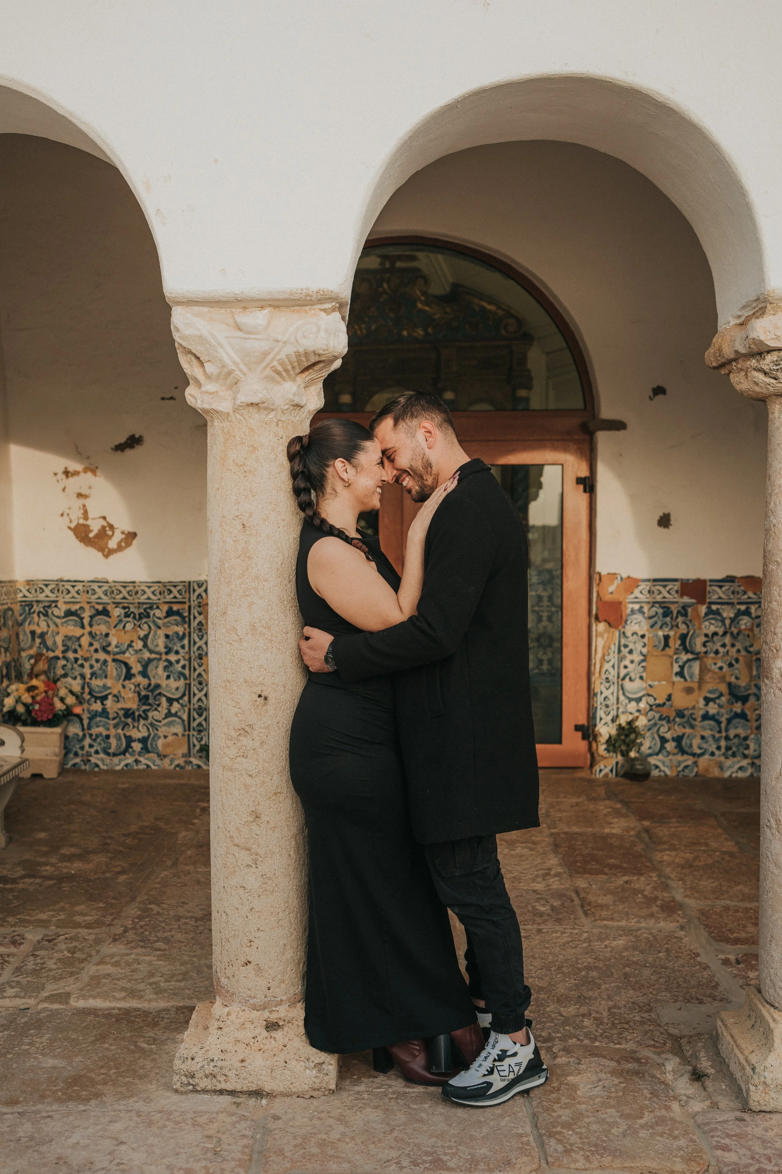 A couple sharing an intimate moment, with foreheads touching, standing under Spanish colonial-style arches in a courtyard with tiled walls and potted plants.