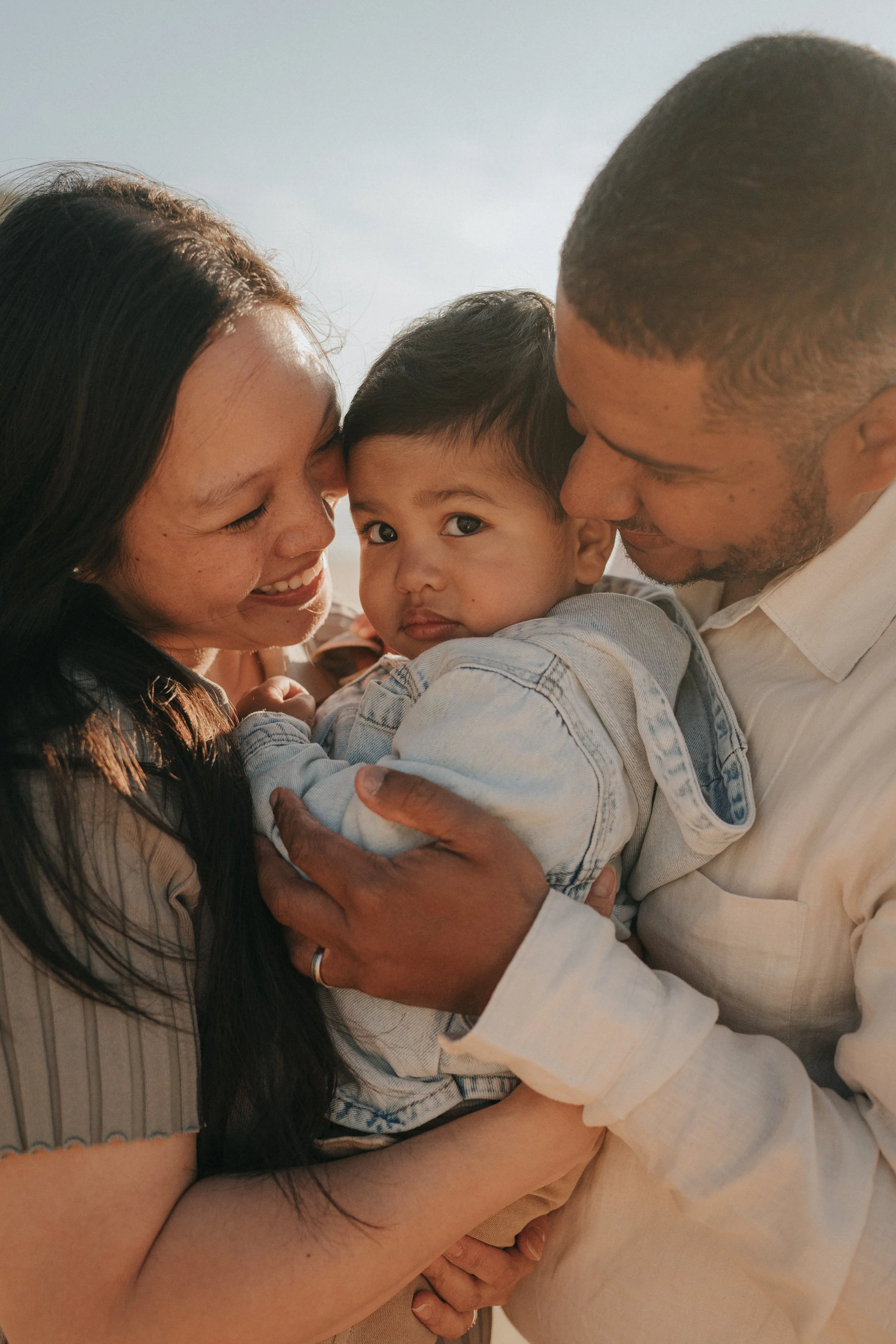 A happy family of three, a woman, a man, and a young child, hugging each other closely and smiling, in natural light.