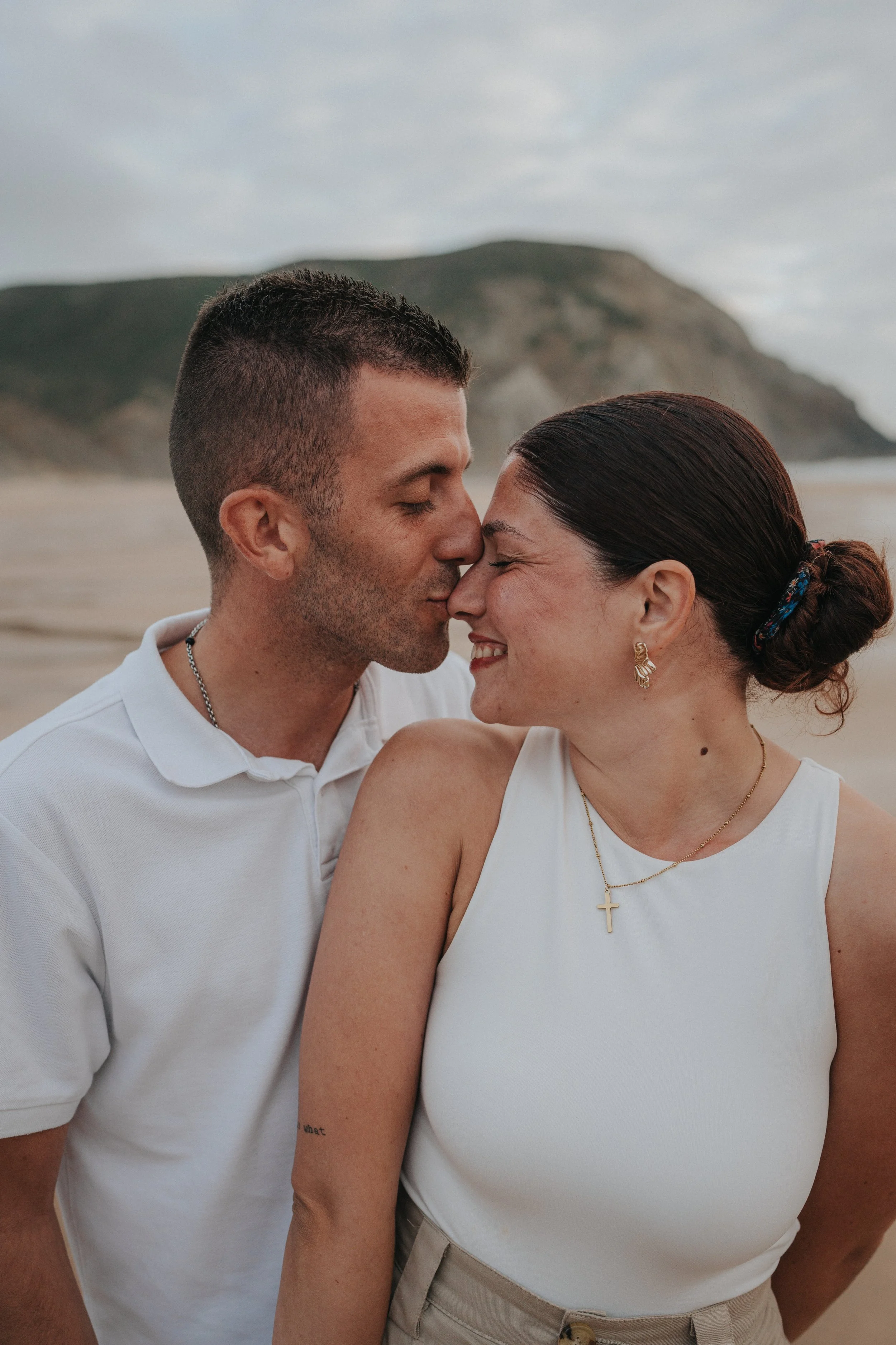 A man and woman are close together with their foreheads touching and eyes closed, smiling on a beach with a mountain in the background.