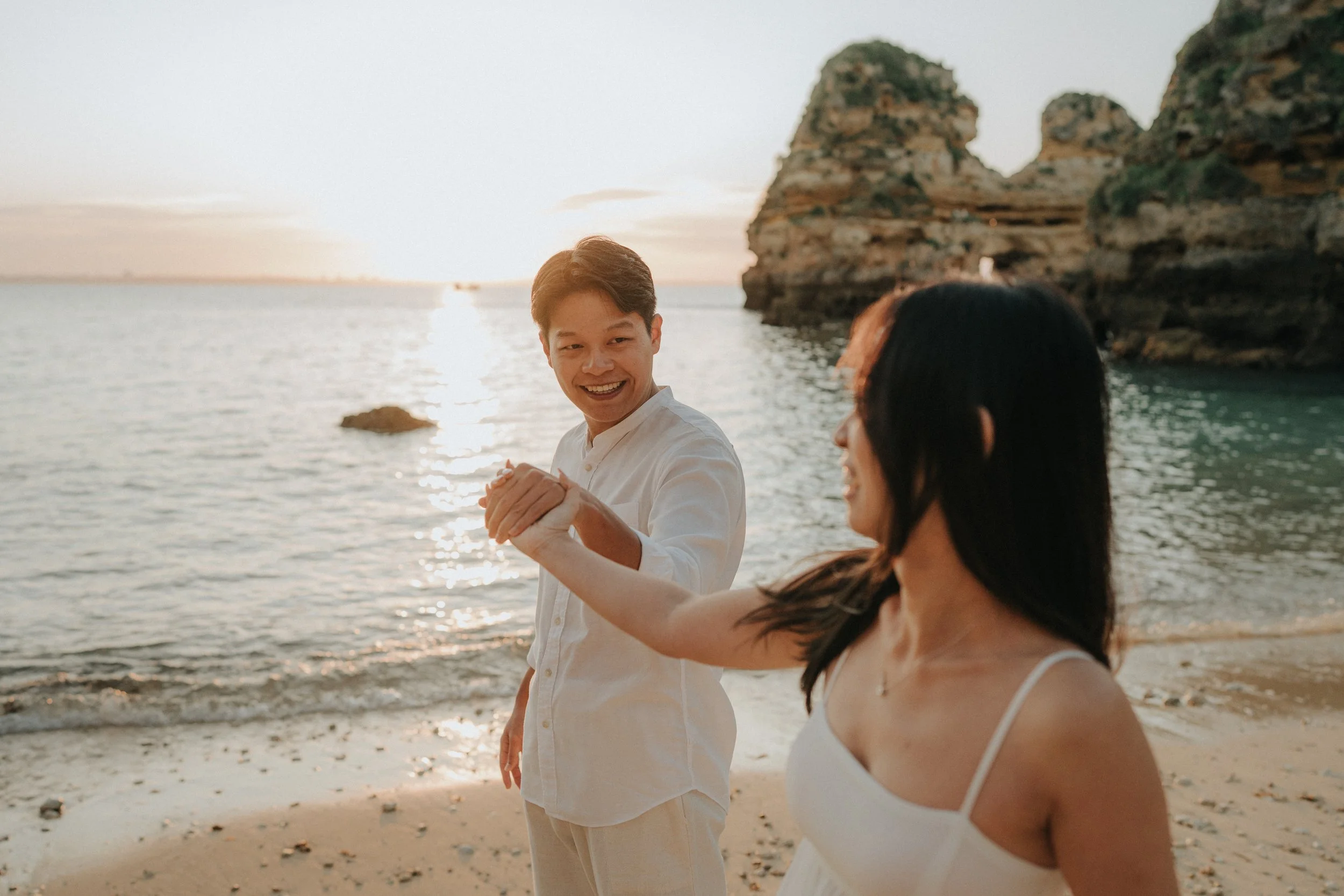 A young man and woman holding hands on a beach at sunset, with large rock formations in the background.
