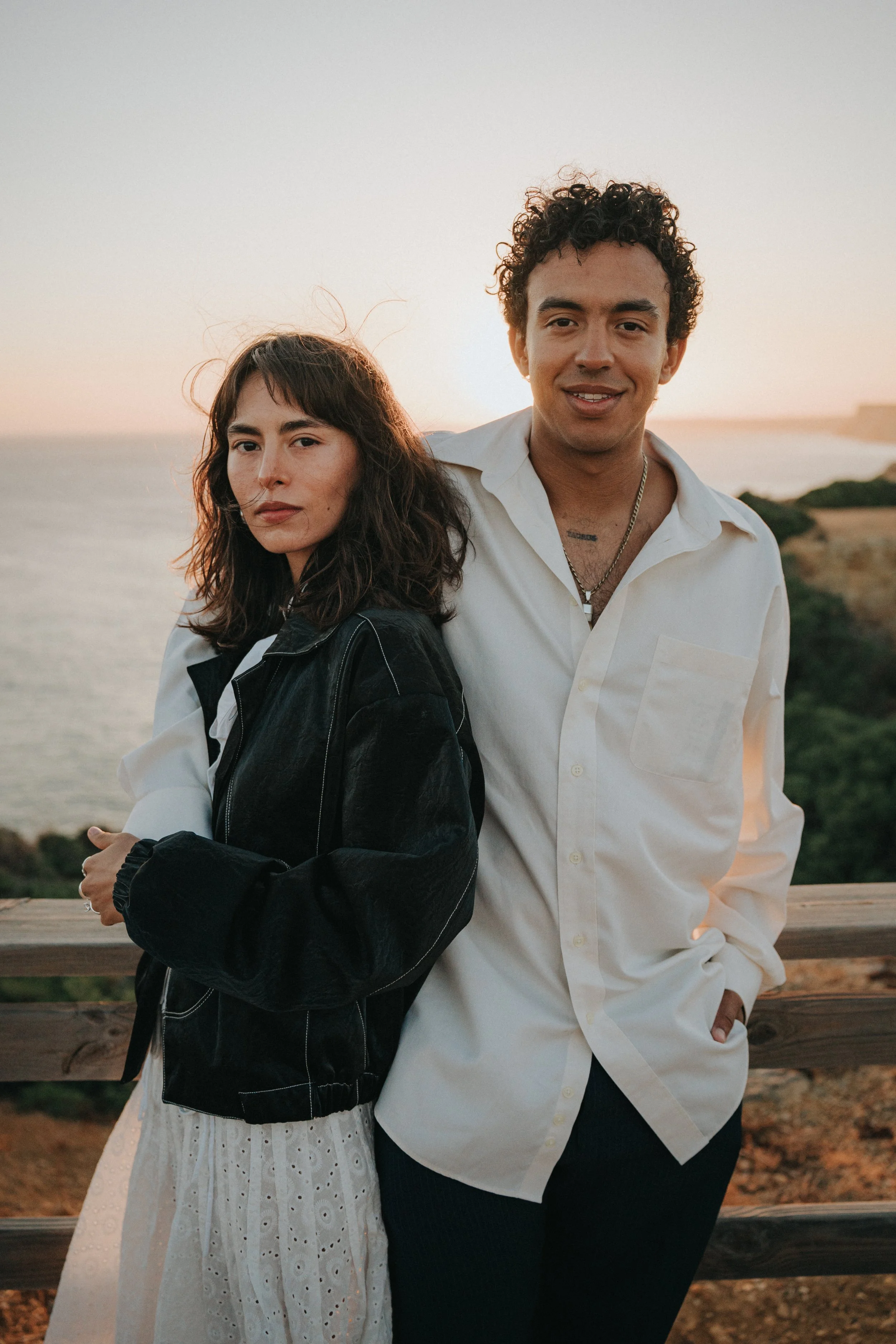 A man and woman stand close together by a wooden railing during sunset on a coastal cliff, with the ocean and sky in the background.