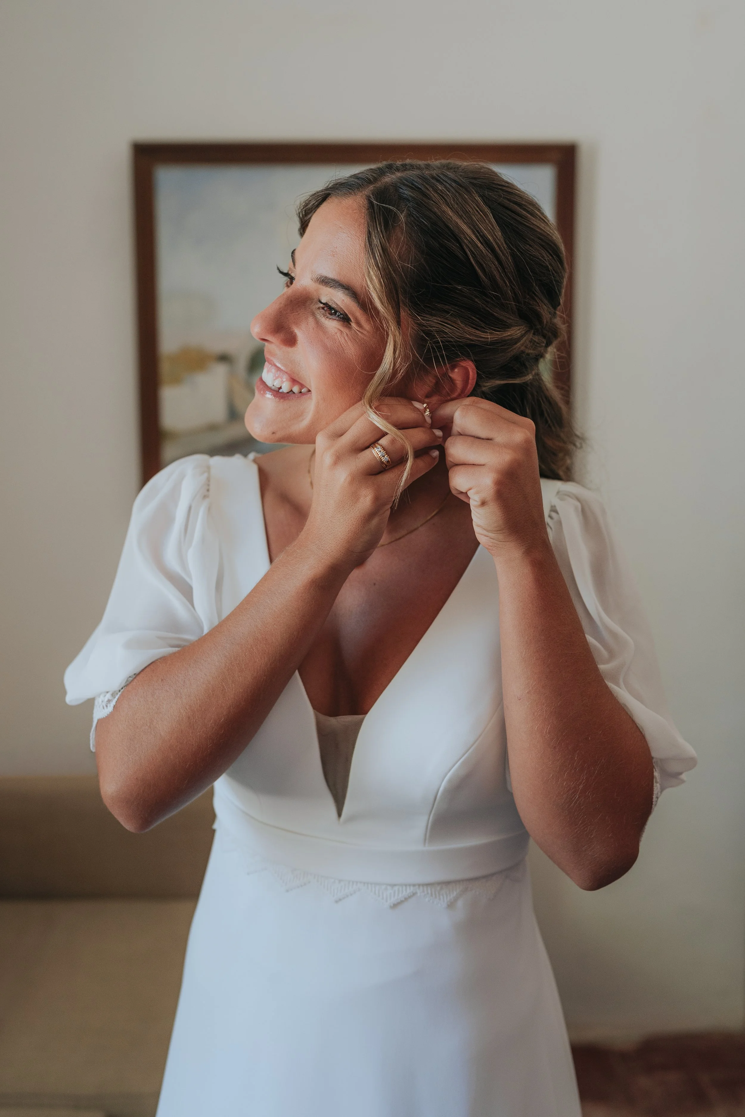 A woman in a white dress with lace details adjusts her earrings, smiling as she prepares for a special event.